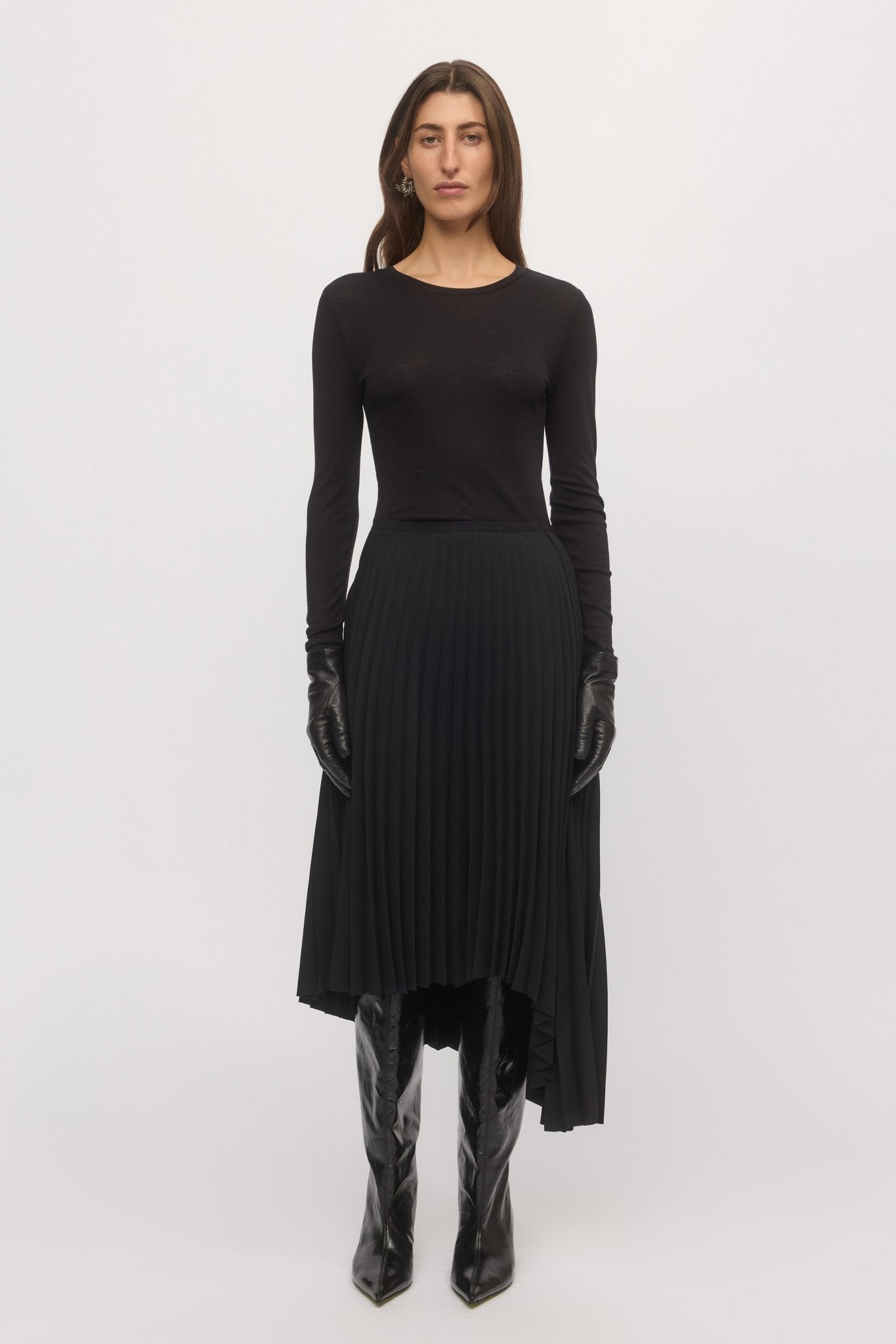 Woman wearing the Asymmetric Pleat Skirt in black – front view of midi-length pleated skirt with long-sleeve black top and boots.