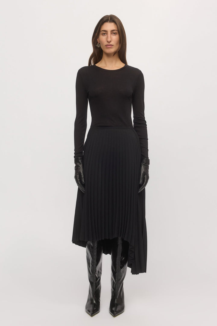 Woman wearing the Asymmetric Pleat Skirt in black – front view of midi-length pleated skirt with long-sleeve black top and boots.