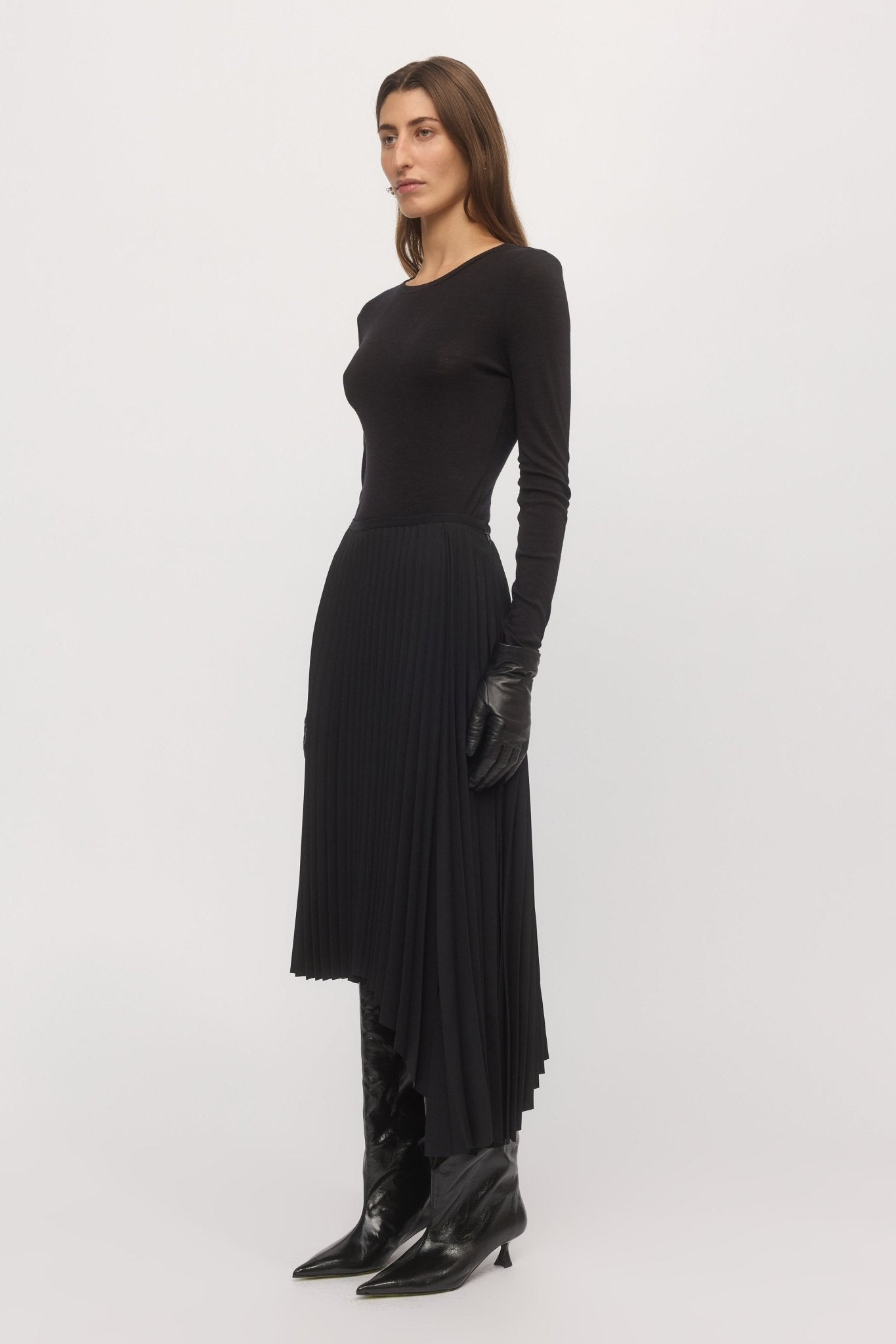 Woman in angled front view wearing black Asymmetric Pleat Skirt with fitted top and knee-high boots.