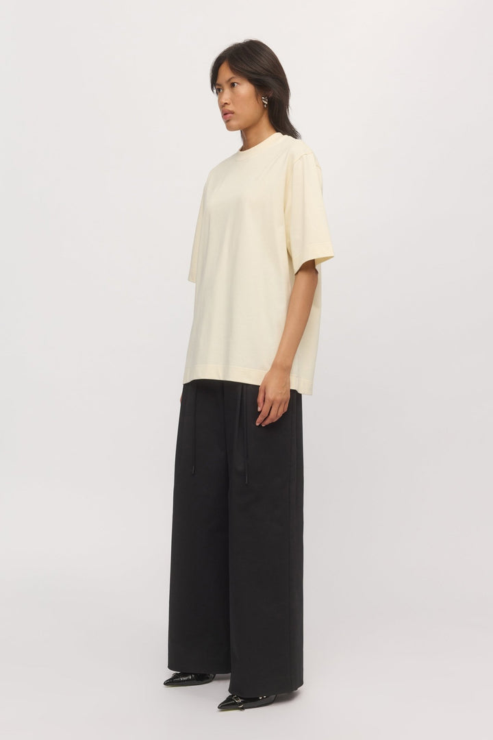 Side view of a woman in the oversized Boyfriend Tee Natural, showing extended short sleeves and a soft drape over wide-leg black pants.