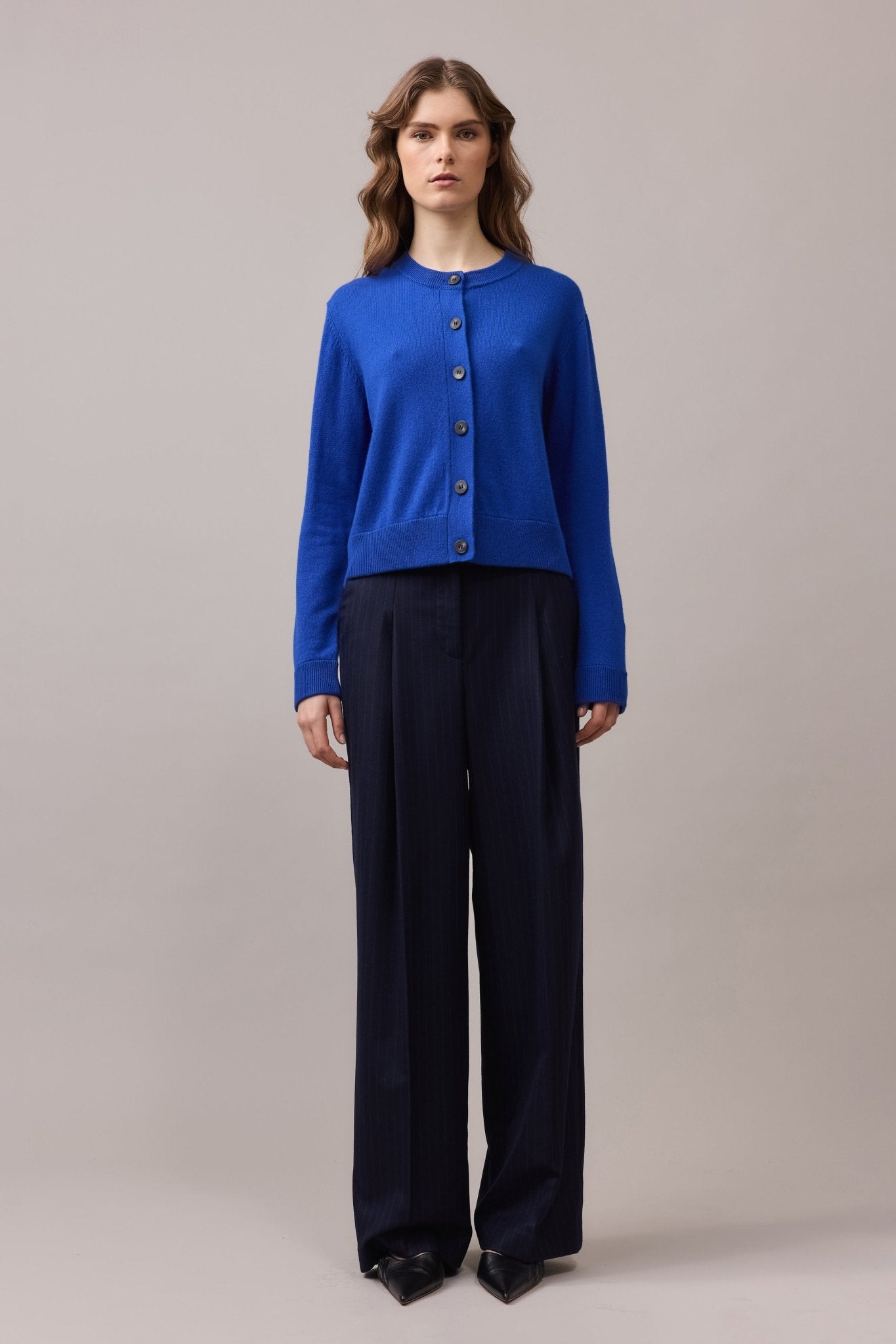 Front view of a woman in a cobalt blue cashmere cardigan with long sleeves and contrast buttons, paired with high-waisted navy trousers.