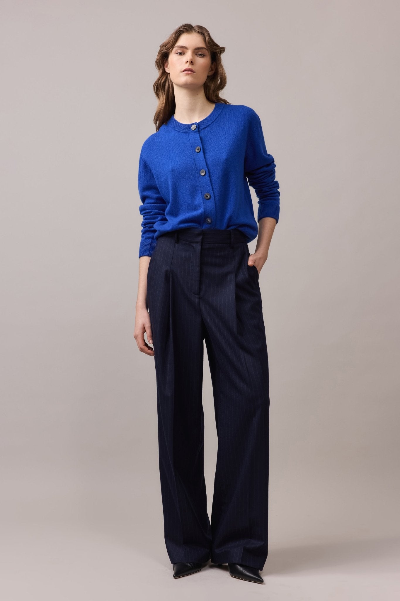 Woman wearing a cobalt blue 100% cashmere crew neck cardigan with mock horn buttons, styled tucked into navy tailored trousers.