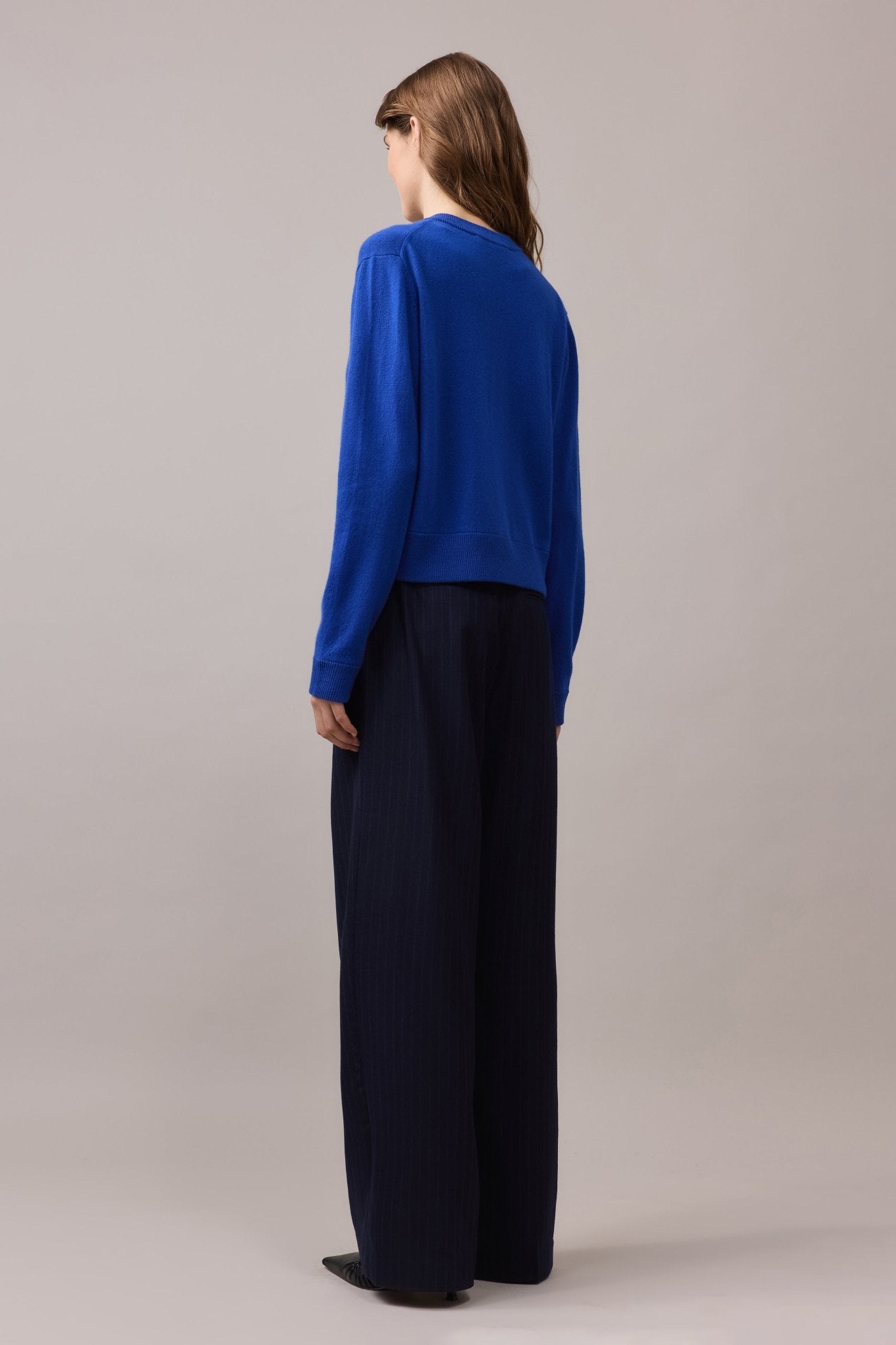 Back view of a woman wearing a cobalt blue cashmere cardigan with a classic crew neck and ribbed hem, paired with navy wide-leg trousers.