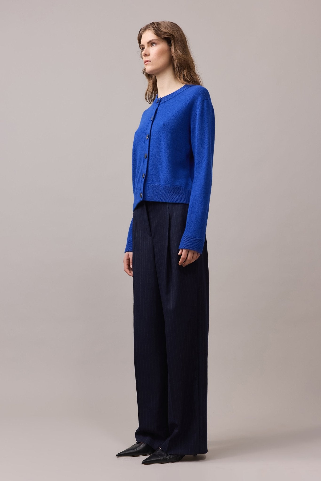 Side view of a woman wearing a cobalt blue crew neck cashmere cardigan with button front, styled with dark wide-leg trousers and pointed shoes.