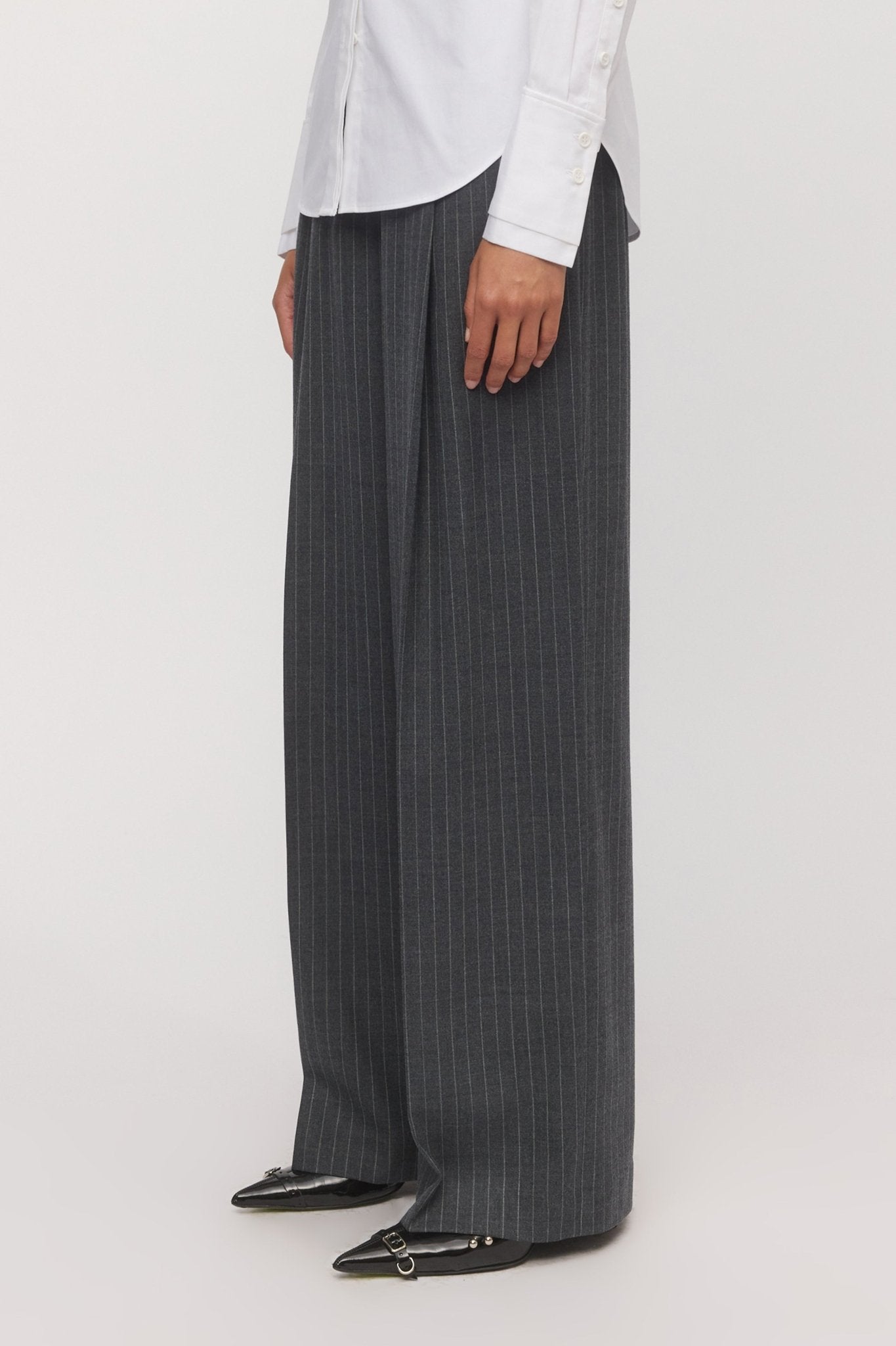 Side view of a woman wearing the Lola Wide Leg Pant in graphite pinstripe, paired with a white shirt and black pointed-toe flats. - Skin and Threads