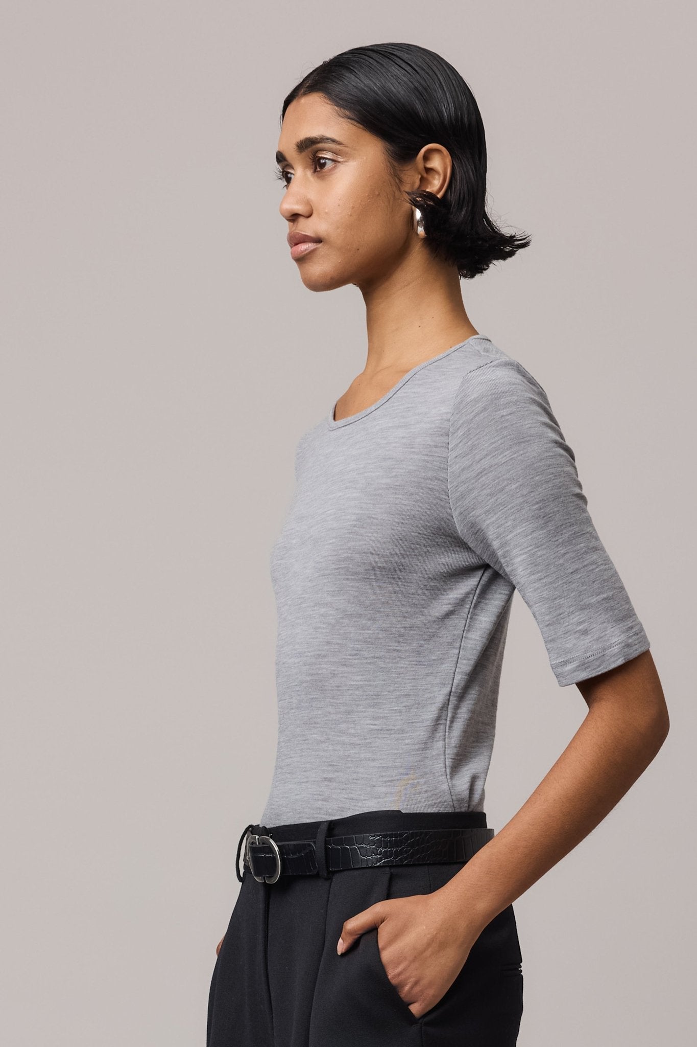 Side close-up of a woman wearing a grey Merino Wool Crew Neck Tee with elbow sleeves and black pants.