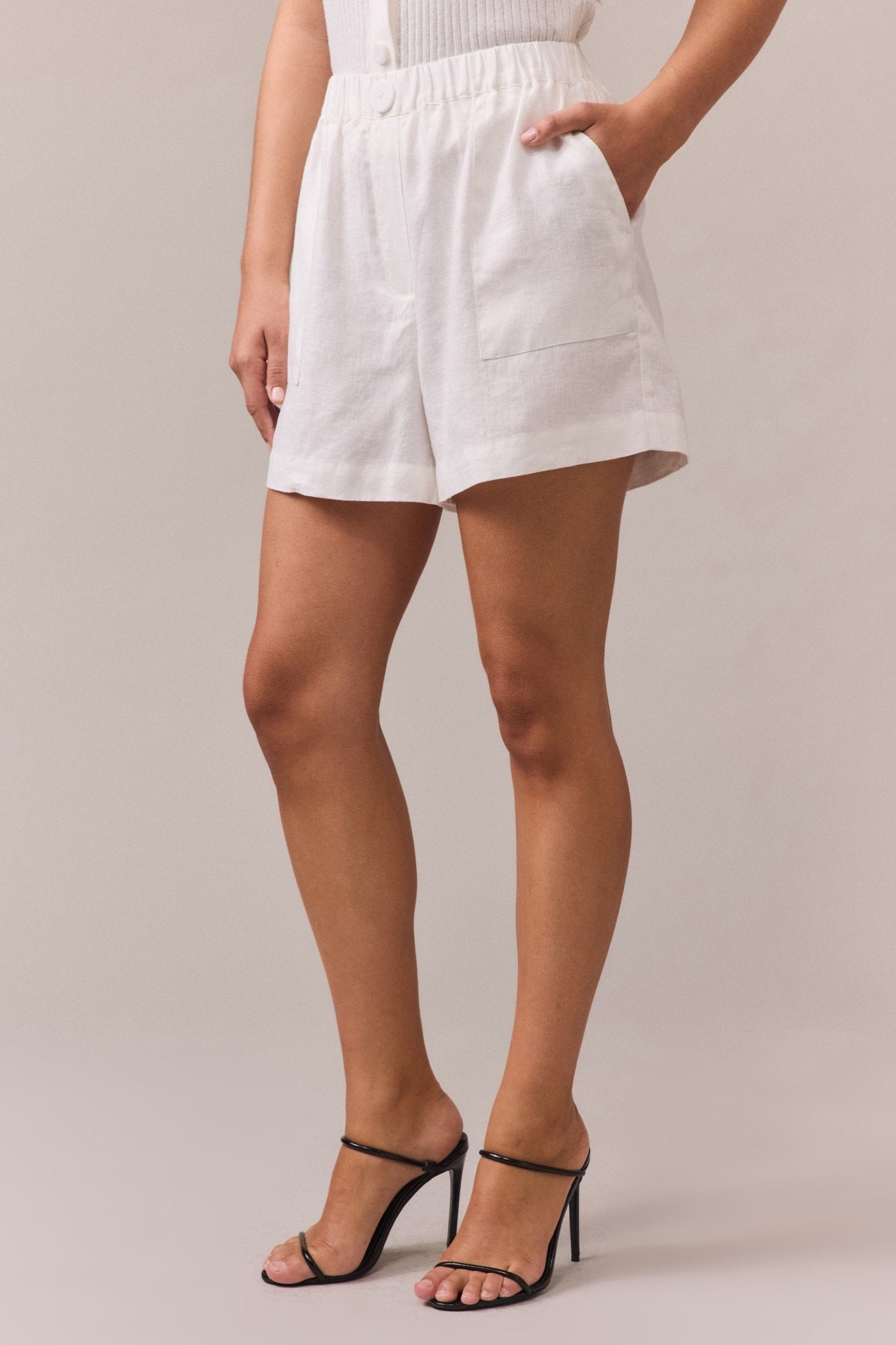 Close-up of a woman wearing white linen shorts with an elastic waistband, feature button, and side seam pockets. Styled with black strappy high heels for an elevated casual look.