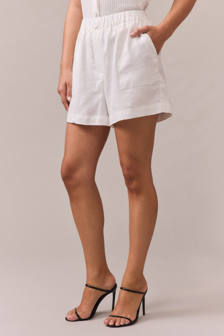 Close-up of a woman wearing white linen shorts with an elastic waistband, feature button, and side seam pockets. Styled with black strappy high heels for an elevated casual look.