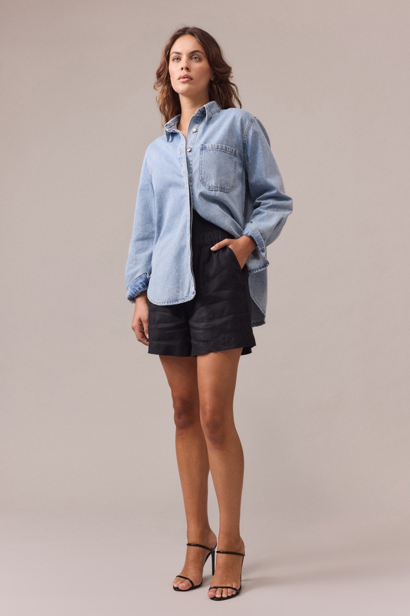 A woman is wearing black Soma linen shorts with one hand in the shorts' pocket, paired with a denim shirt.