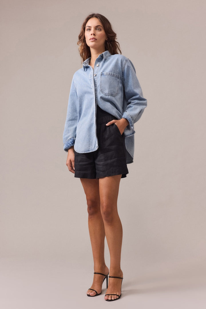 A woman is wearing black Soma linen shorts with one hand in the shorts' pocket, paired with a denim shirt.