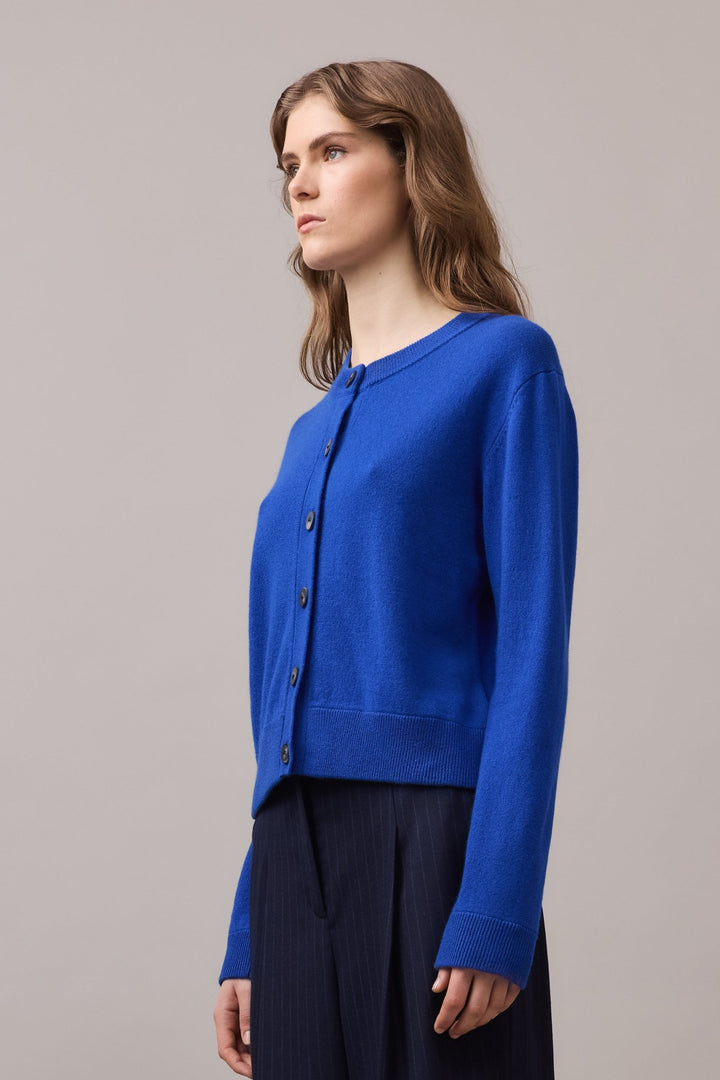 Side angle of a woman wearing a cobalt blue cashmere cardigan, showing the relaxed fit and mock horn button detailing from the front.