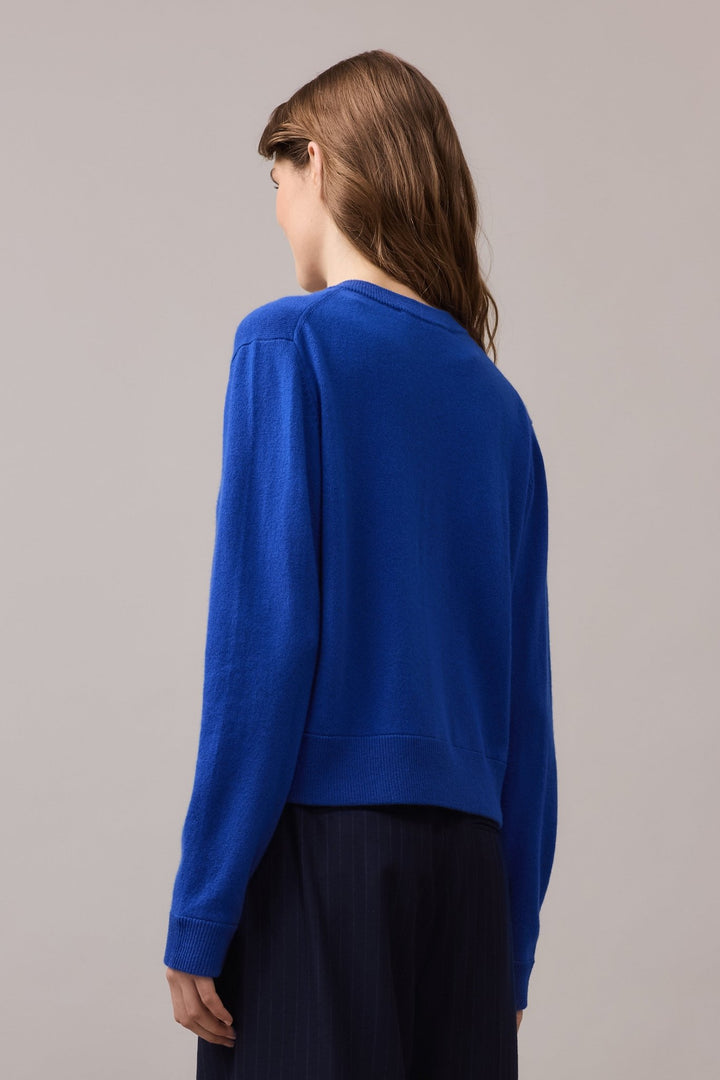 Back view of a woman wearing a cobalt blue 100% cashmere crew neck cardigan, showing the ribbed hem and soft drape over tailored trousers.
