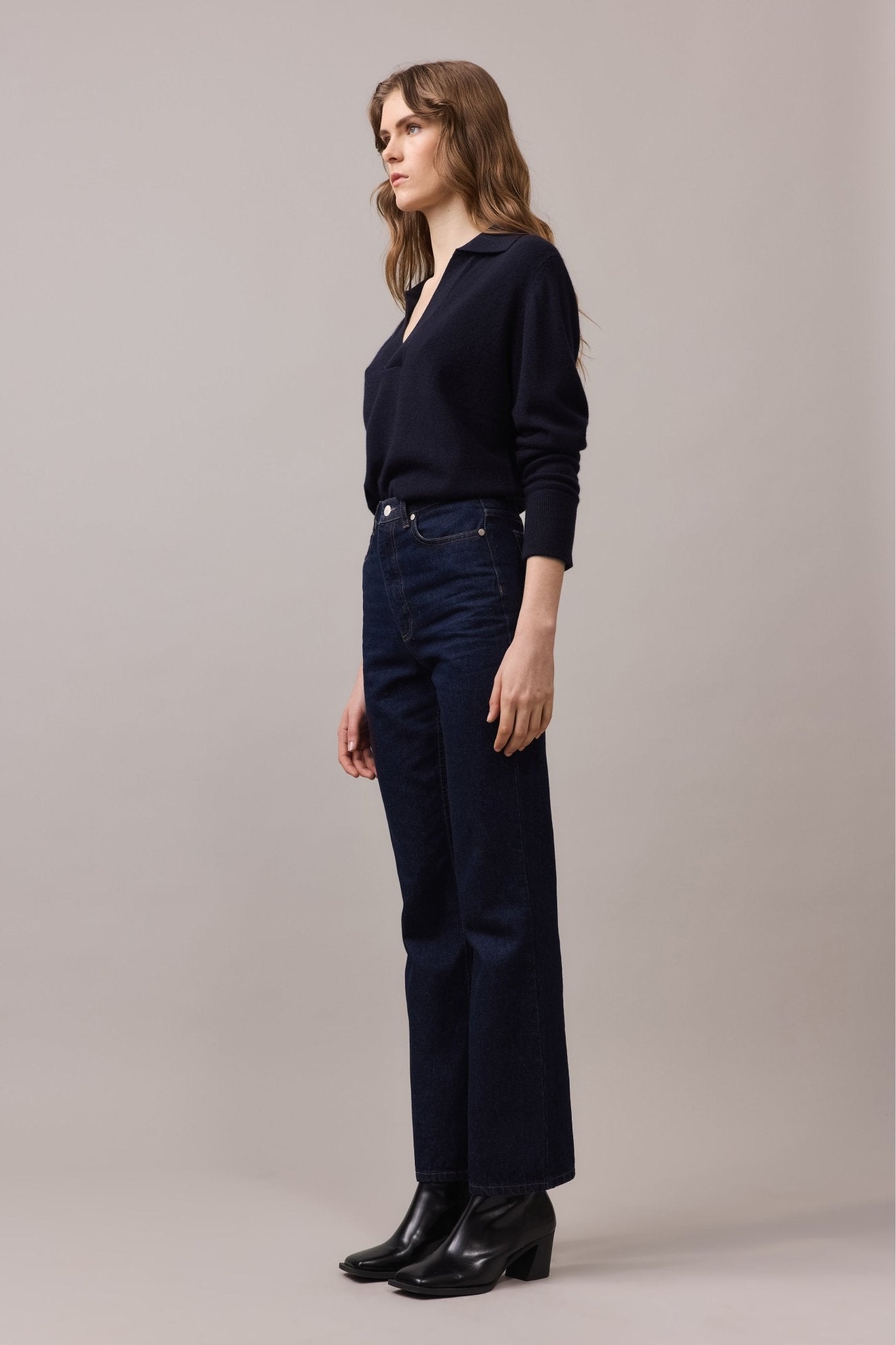 Side view of a woman wearing the Cashmere Polo Knit in 100% cashmere, paired with dark blue high-waist jeans. The V-neck with collar and long sleeves create a relaxed, stylish look