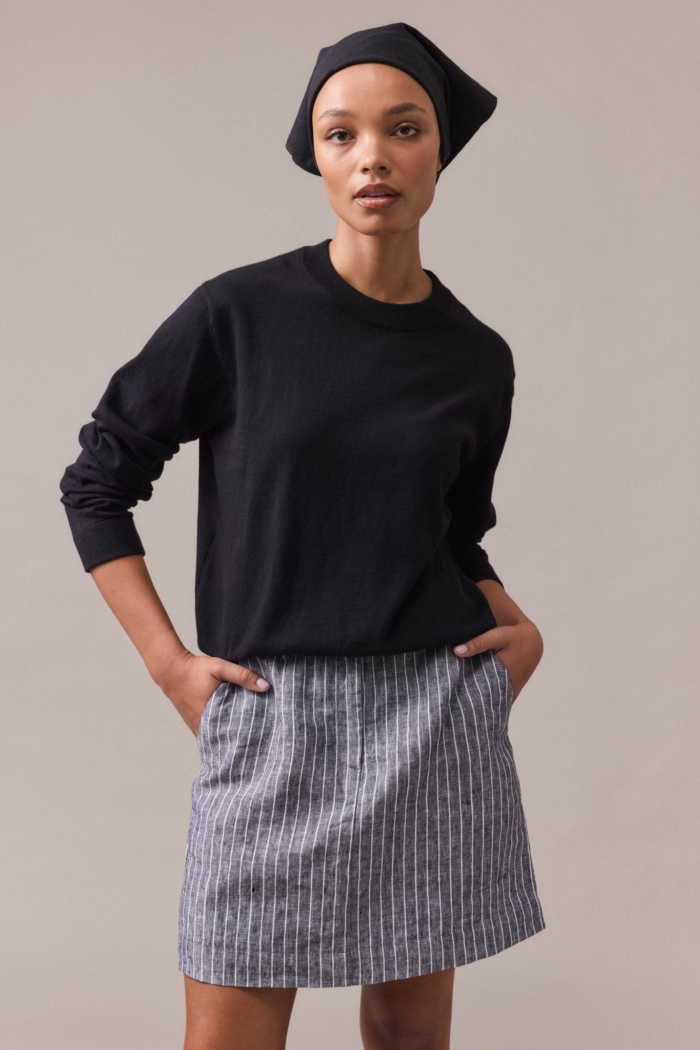 A woman wearing a black cotton-cashmere crew neck knit paired with a grey skirt, with her hands in the pockets.