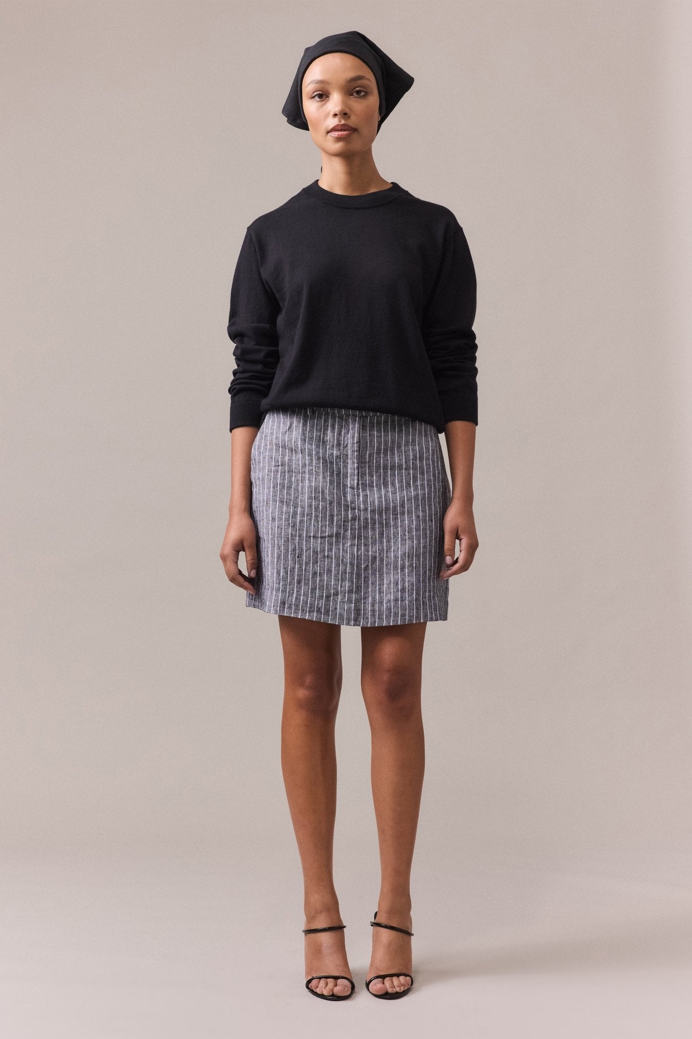 A woman wearing a black cotton-cashmere crew neck knit paired with a grey skirt, standing with a relaxed posture.