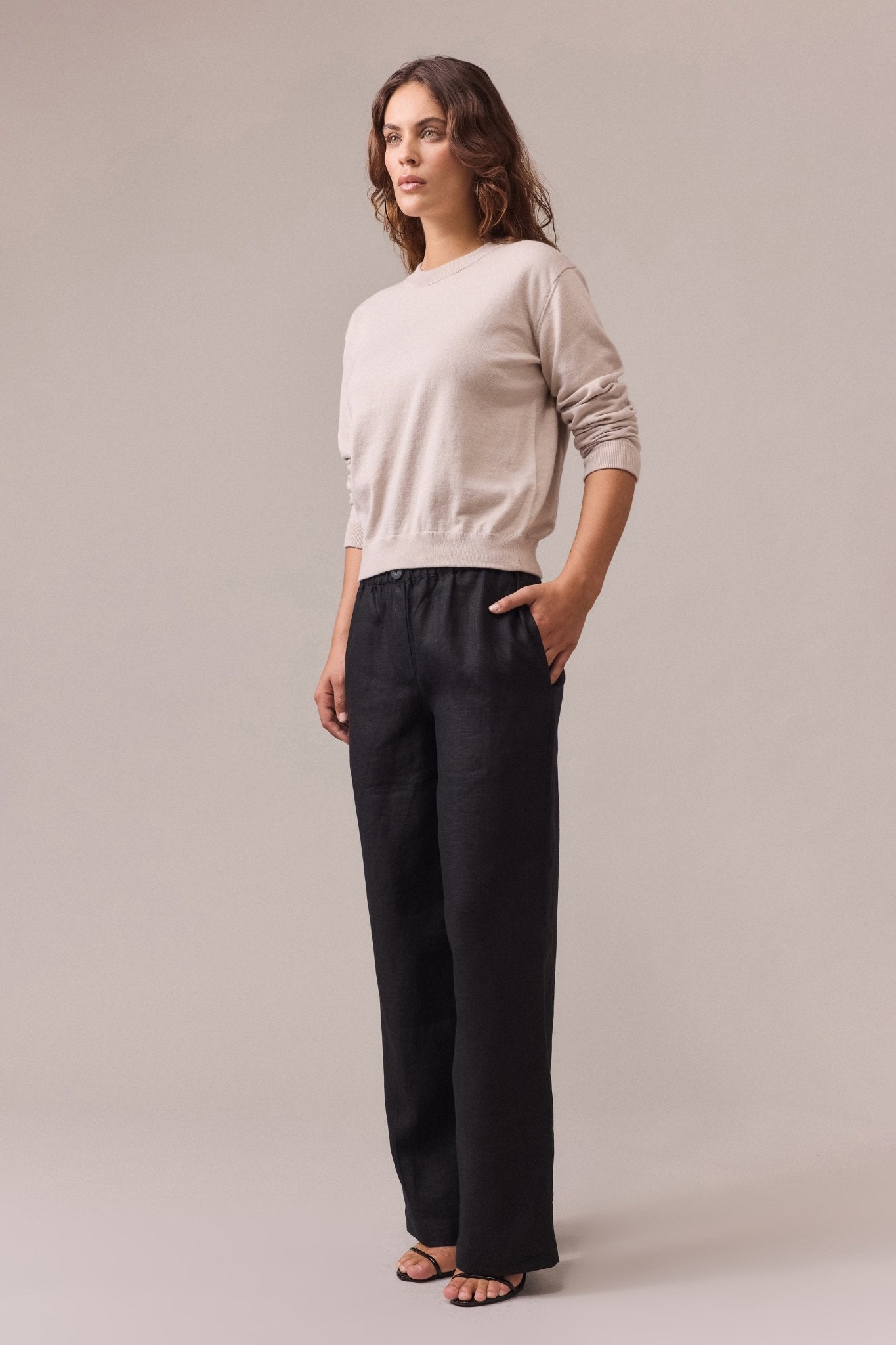 A woman wearing a sand cotton-cashmere crew neck knit with long sleeves, paired with long wide-leg black pants and her hand in the pocket.