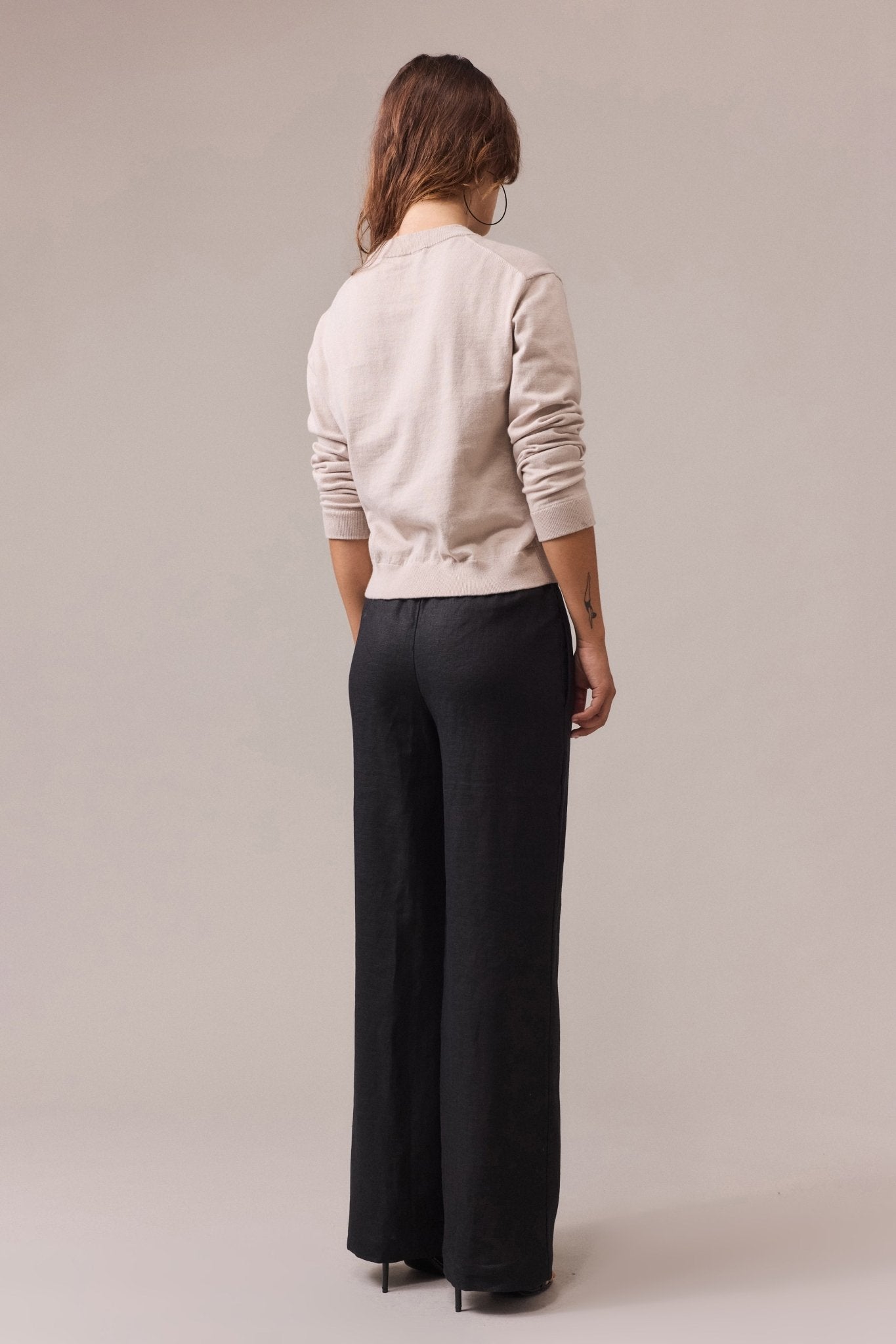 A woman seen from the back, wearing a sand cotton-cashmere crew neck knit with long sleeves, paired with long wide-leg black pants.