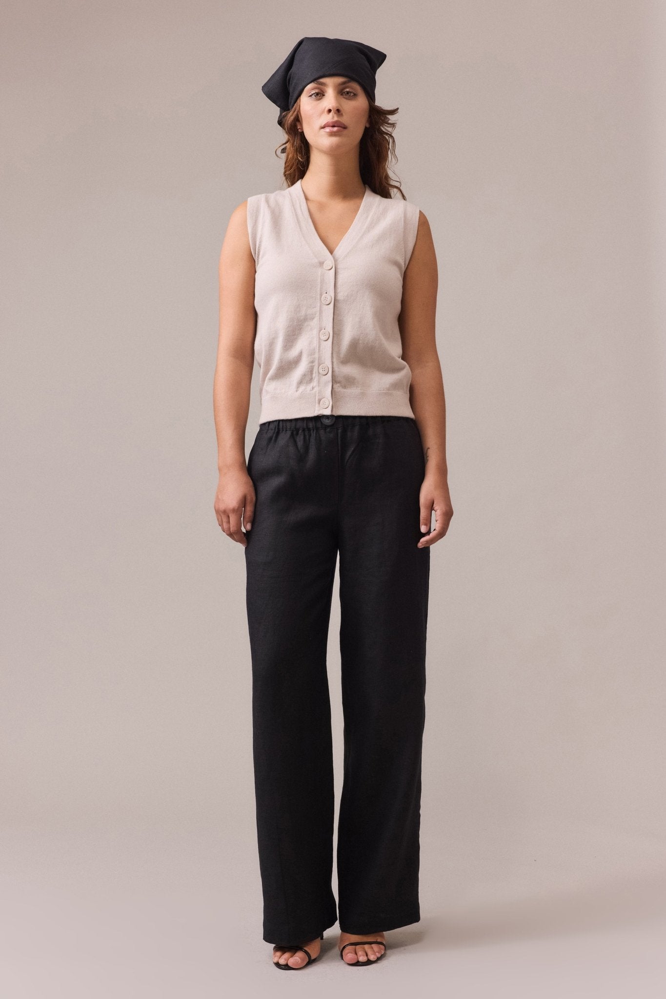 A woman wearing a sand button-up cotton cashmere vest, paired with a wide leg black linen pant.