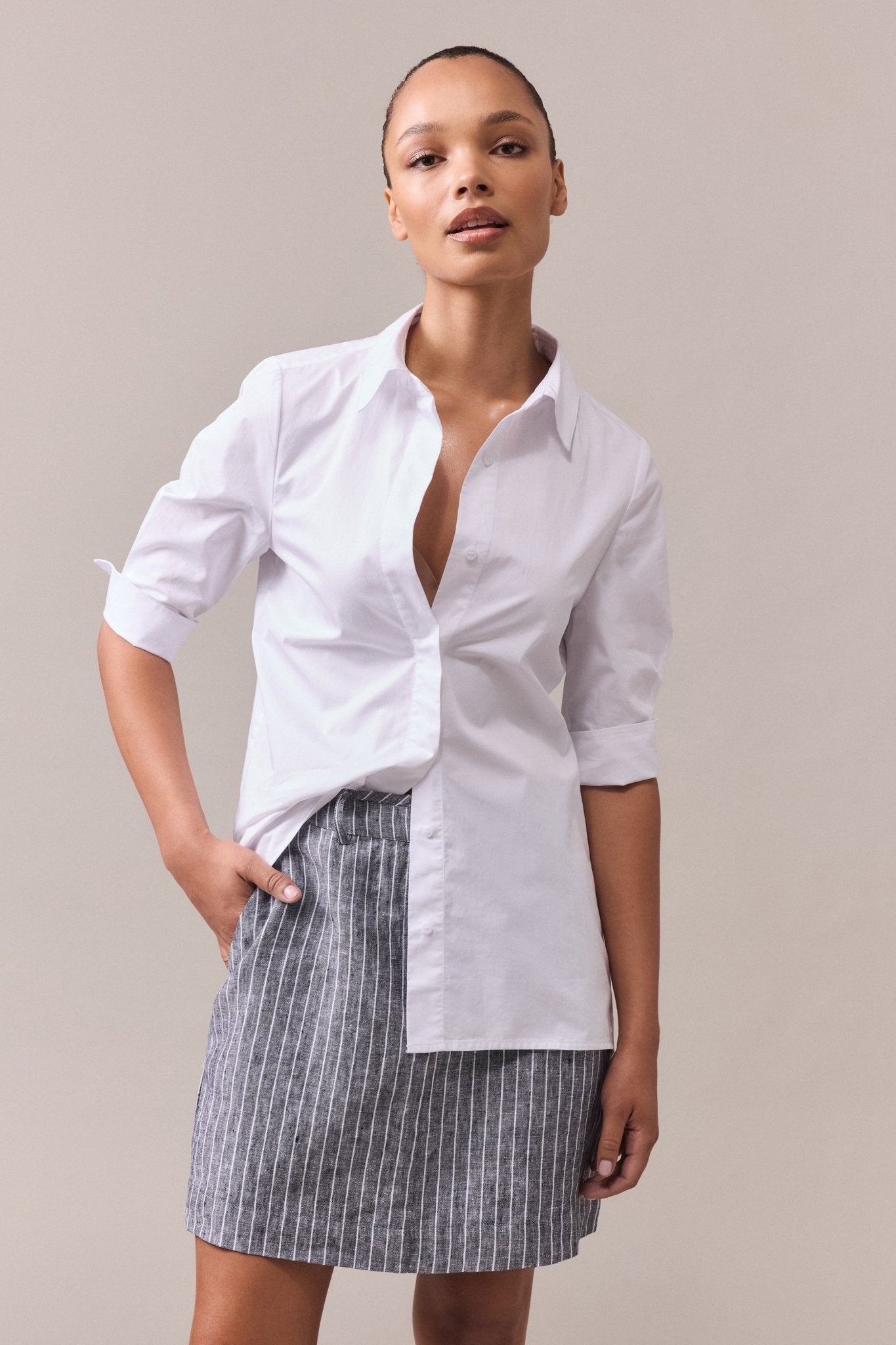 Woman wearing white elbow sleeve cotton shirt, with one hand in the skirt pocket and unbuttoned details revealing a relaxed styling approach.