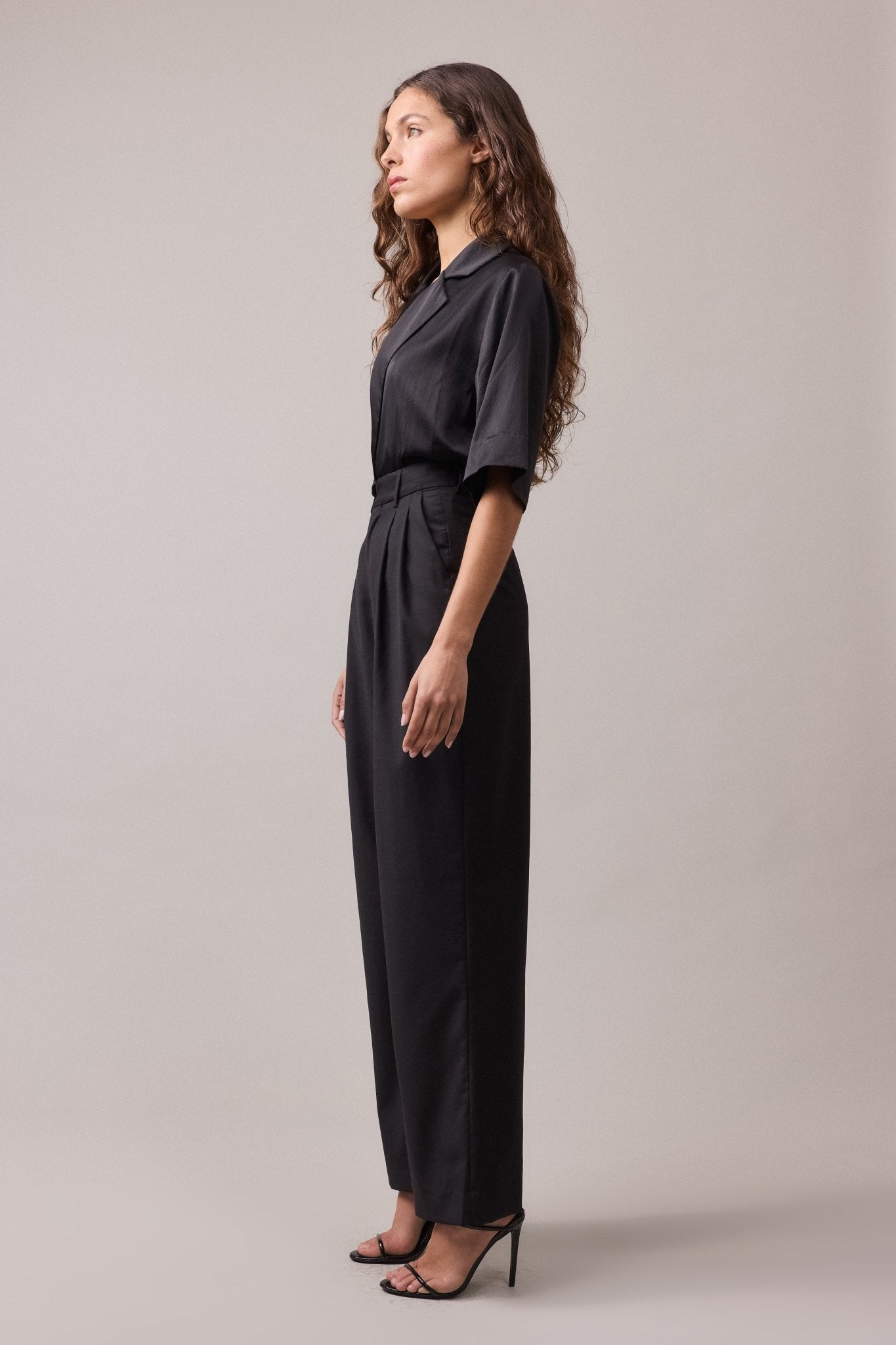 Side view of the woman wearing the black Frankie Wide Leg Pant in wool, paired with a black shirt. The high-waist, wide-leg fit and relaxed silhouette are showcased, creating a flattering, stylish look.