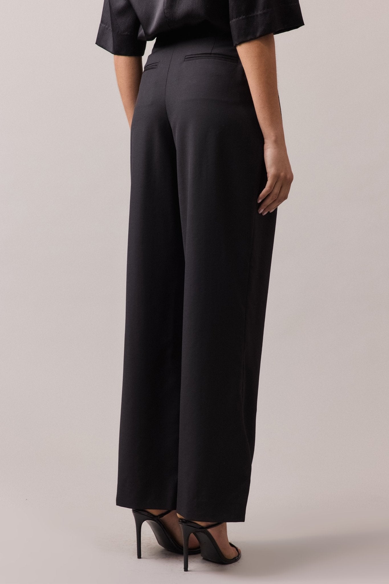 View of the woman from behind, wearing the black Frankie Wide Leg Pant in 100% wool. The wide-leg, high-waist fit is visible, along with the back jet pockets.