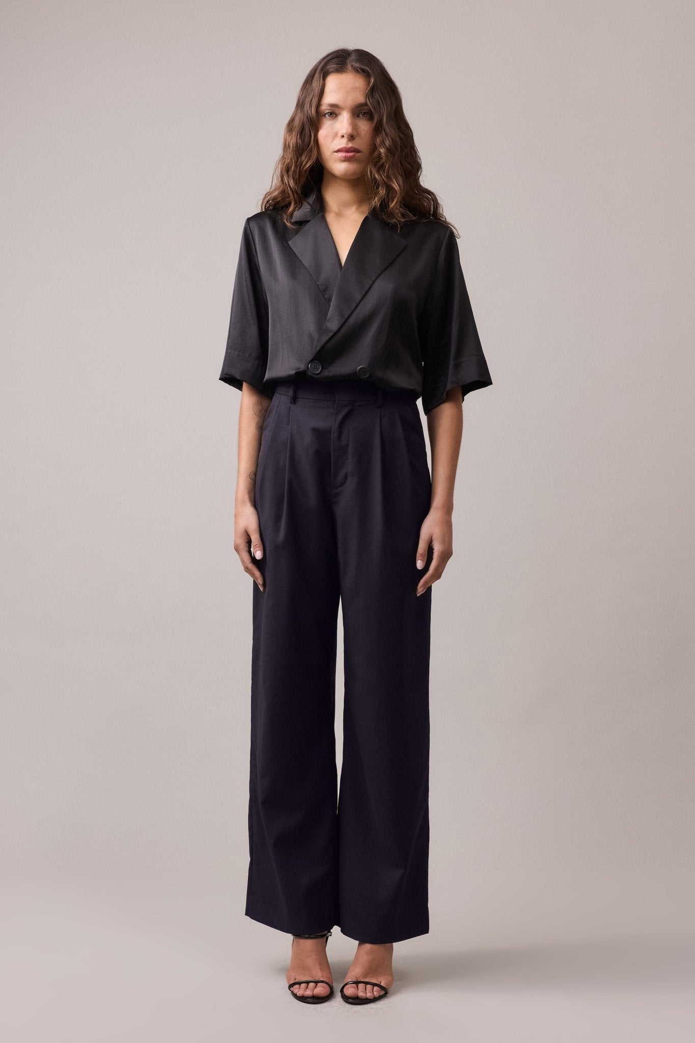 Woman wearing the Frankie Wide Leg Pant in wool, paired with a black shirt. The high-waist, wide-leg fit of the pants is complemented by the relaxed silhouette, showcasing the stylish and flattering combination.