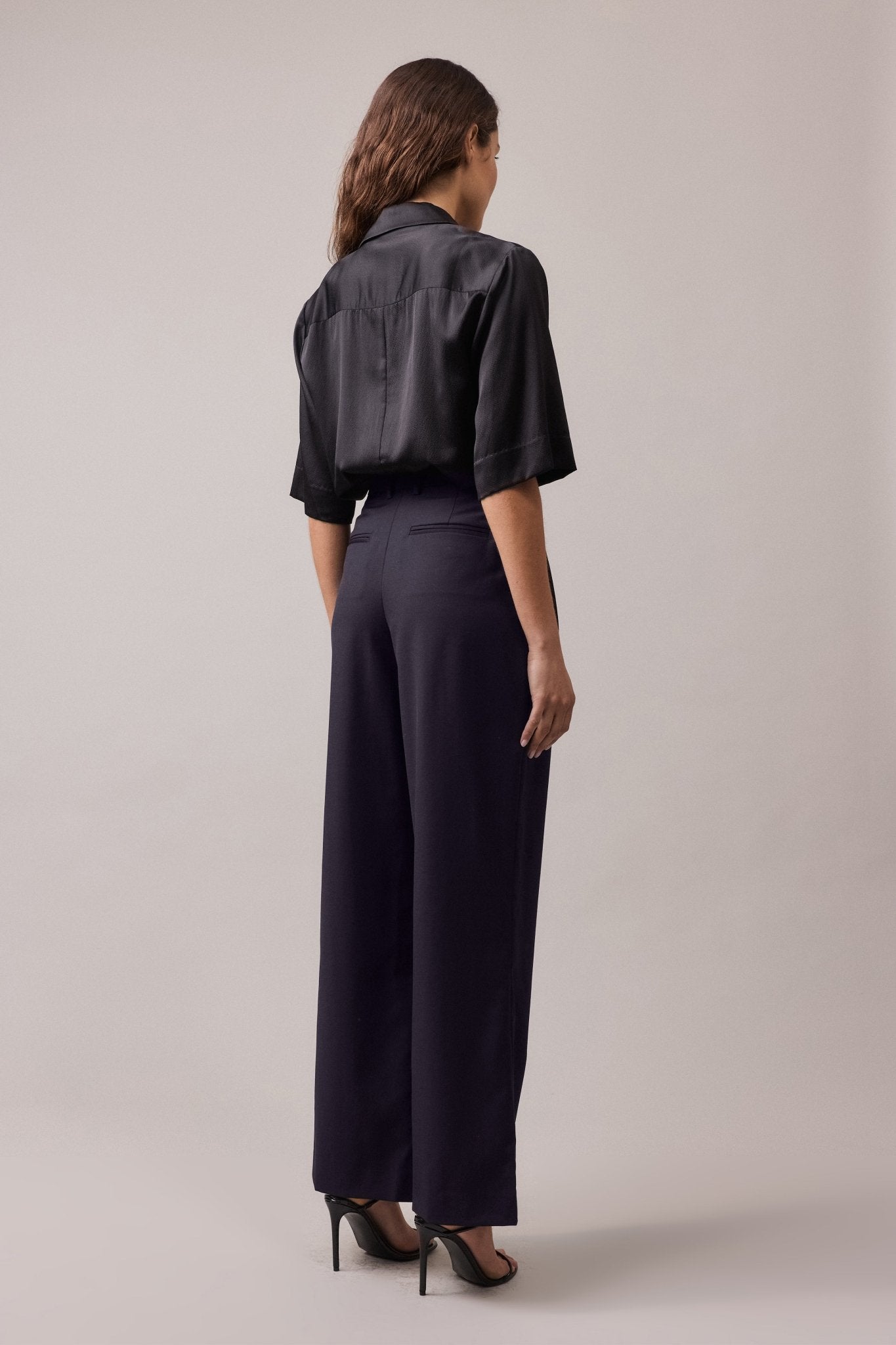 View of the woman from behind, wearing the Frankie Wide Leg Pant in 100% wool, paired with a black shirt. The high-waist, wide-leg fit and back jet pockets are visible, creating a flattering and relaxed silhouette.