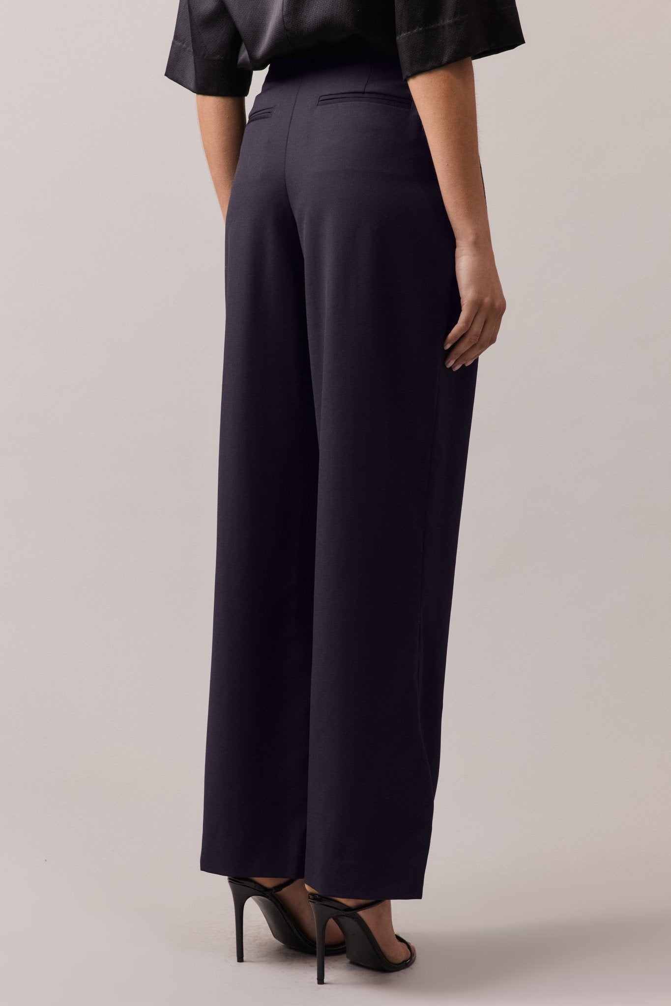 View of the woman from behind, wearing the Frankie Wide Leg wool pant. The wide-leg, high-waist fit is visible, along with the back jet pockets, showcasing the flattering and relaxed silhouette.