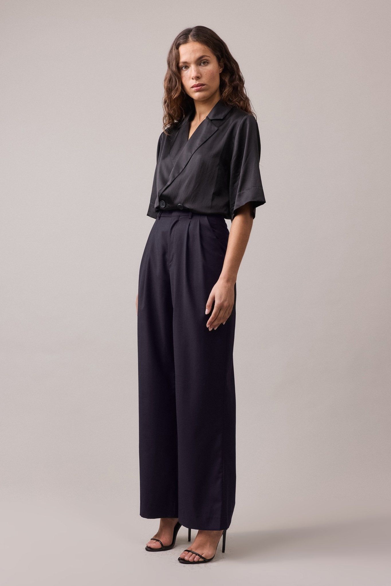 Full view of the woman wearing the Frankie Wide Leg Pant in wool, paired with a black shirt. The wide-leg, high-waist fit of the pants creates a relaxed, flattering silhouette, offering a stylish and versatile look.