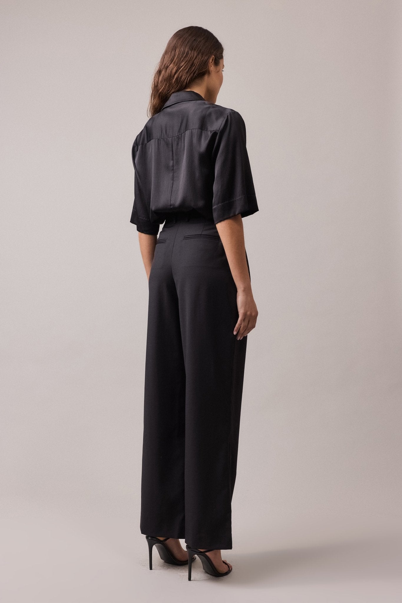 View of the woman from behind, wearing the black Frankie Wide Leg Pant in 100% wool, paired with a black shirt. The high-waist, wide-leg fit and back jet pockets are visible.