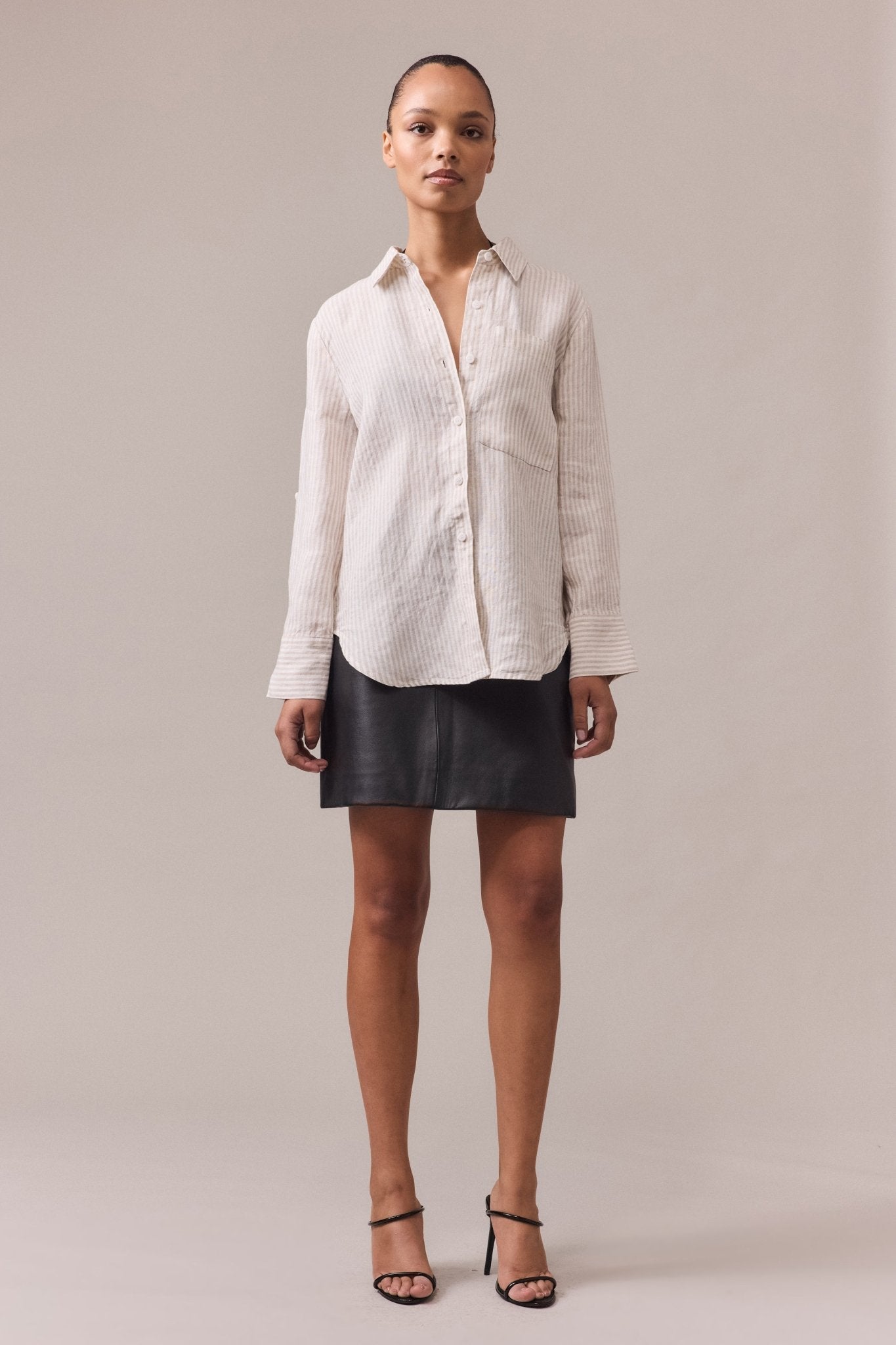 A woman wearing a longline white linen button-through shirt with a patch chest pocket and rolled-up sleeves, paired with a leather mini skirt.