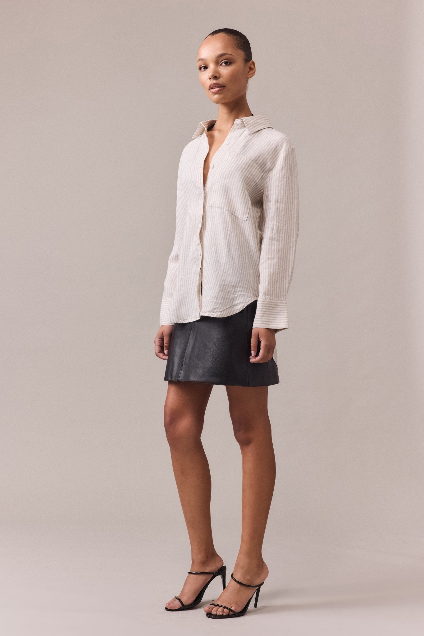 A woman wearing a longline white linen button-through shirt with a patch chest pocket and rolled-up sleeves, styled with a leather mini skirt.