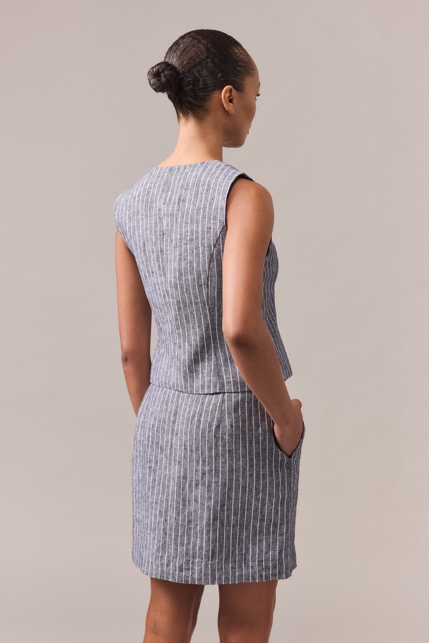 A woman seen from the back, wearing the Kaia stripe linen vest paired with the matching Kaia Stripe Linen Mini Skirt, with her hands in the pockets.