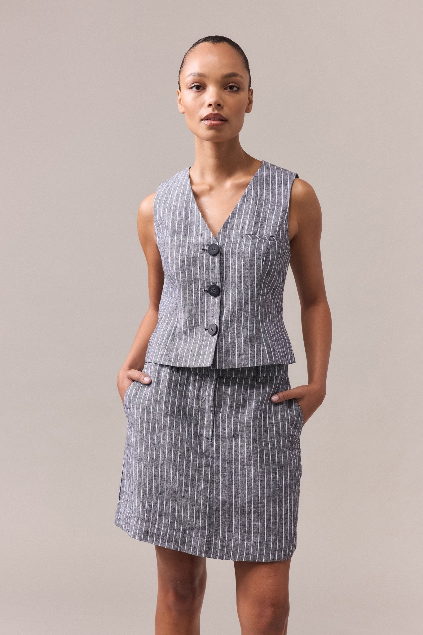 A woman wearing the striped linen vest with princess seams, paired with the matching Kaia Stripe Linen Mini Skirt and her hands in the skirt pockets.