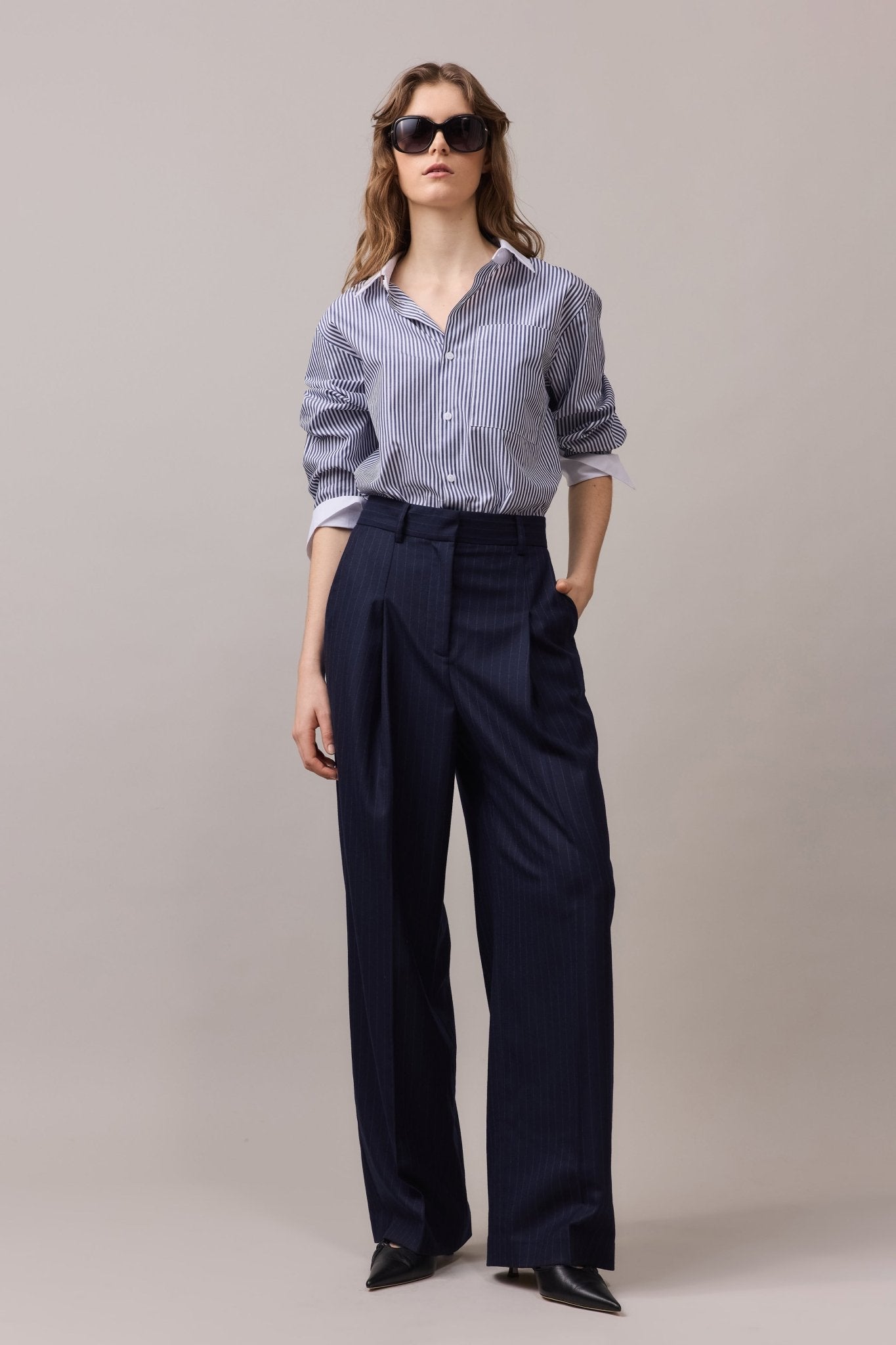 Woman wearing the Lenny Stripe Shirt, a classic relaxed-fit cotton shirt with a contrast collar and cuffs, paired with tailored navy trousers and black pointed-toe shoes. The shirt features a front patch pocket and a clean button-through placket.