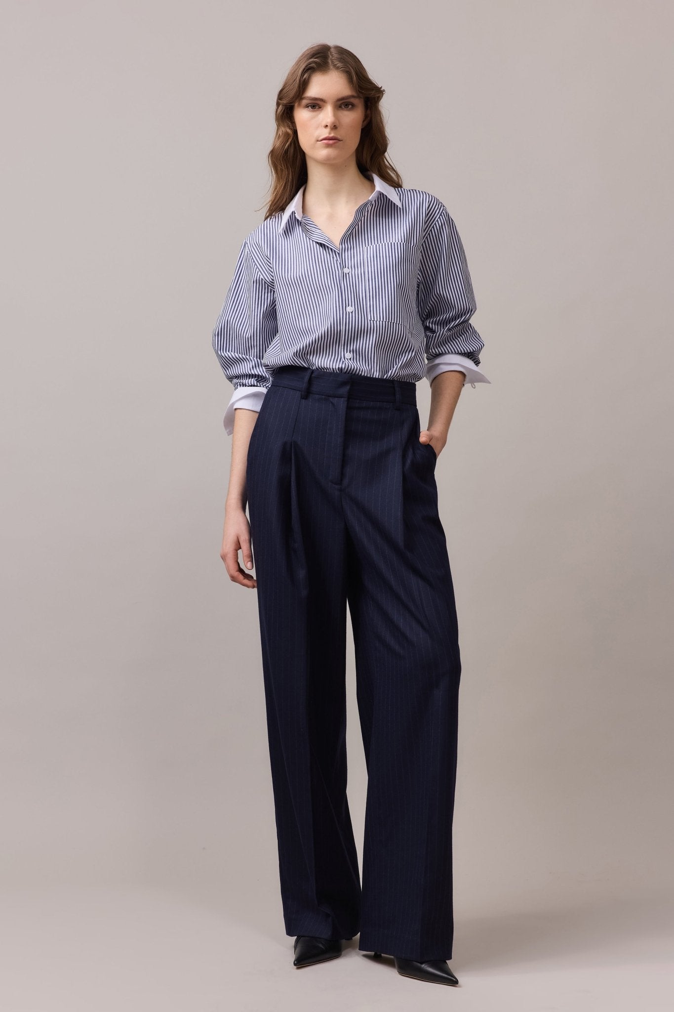 Woman wearing the Lenny Stripe Shirt with sleeves rolled up, paired with tailored navy trousers. One hand is in the pocket, showcasing the relaxed yet classic fit of the outfit.