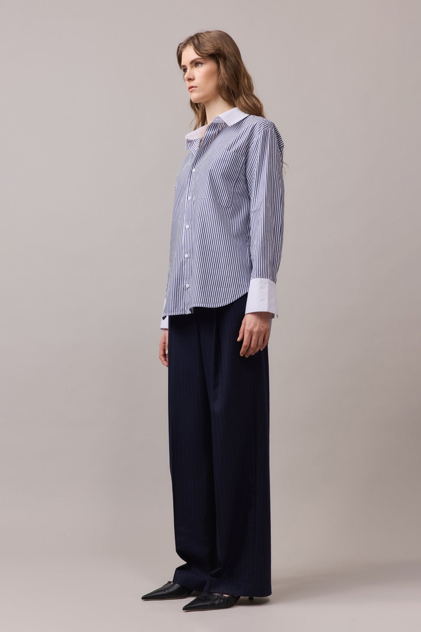 Woman wearing the Lenny Stripe Shirt with sleeves down, revealing the white contrast cuffs and cotton collar. Paired with tailored navy trousers for a polished look.