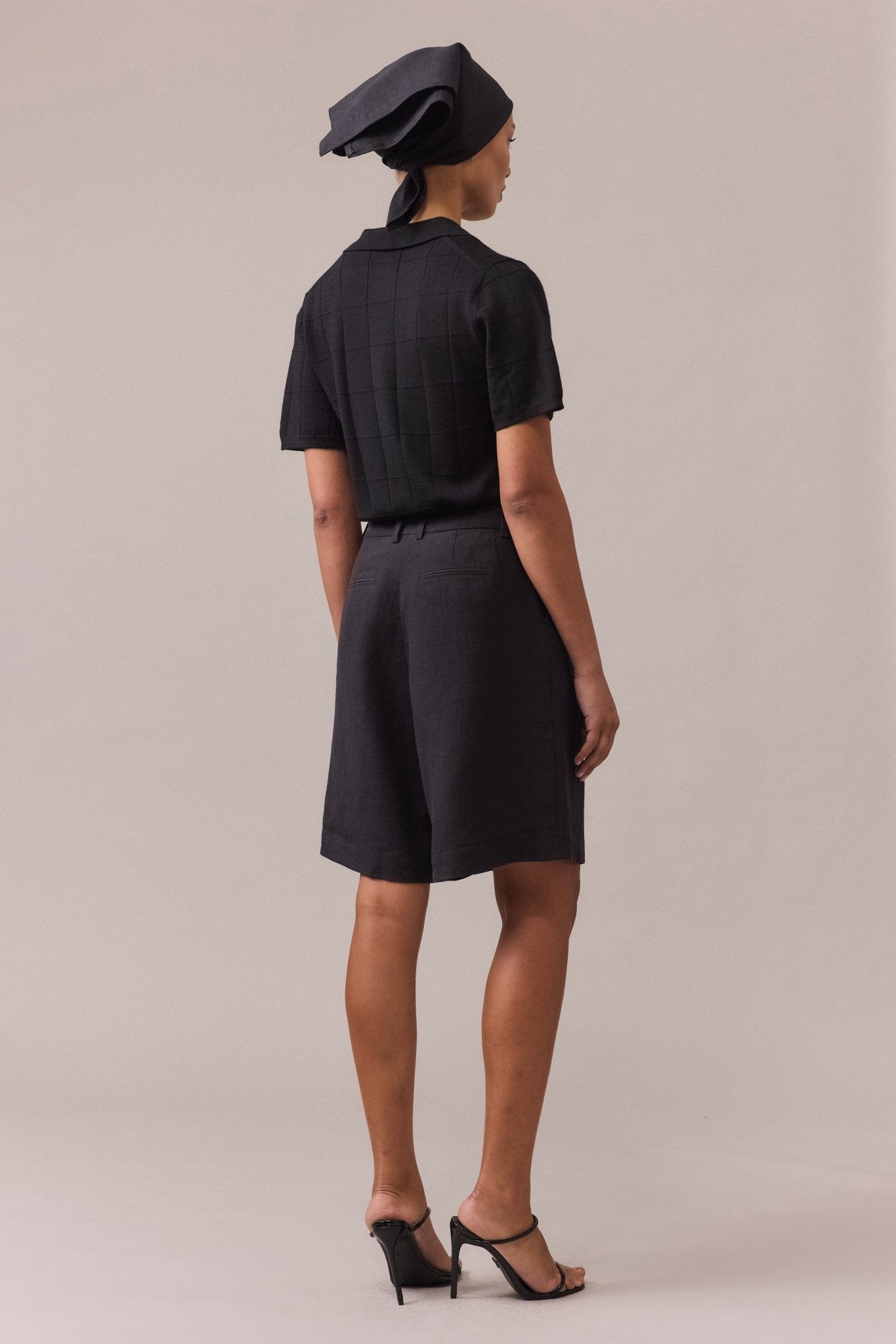 Back view of a woman wearing a black polo knit and black tailored shorts.