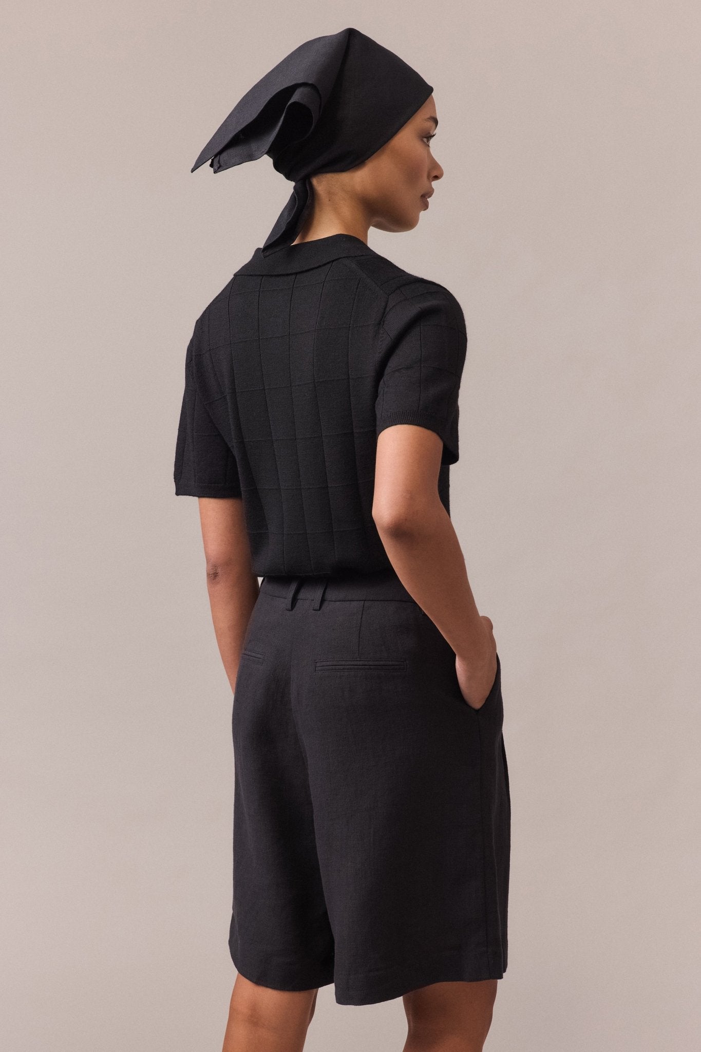 Back view of a woman wearing a black Merino wool polo knit and black tailored shorts, with one hand in the pocket.