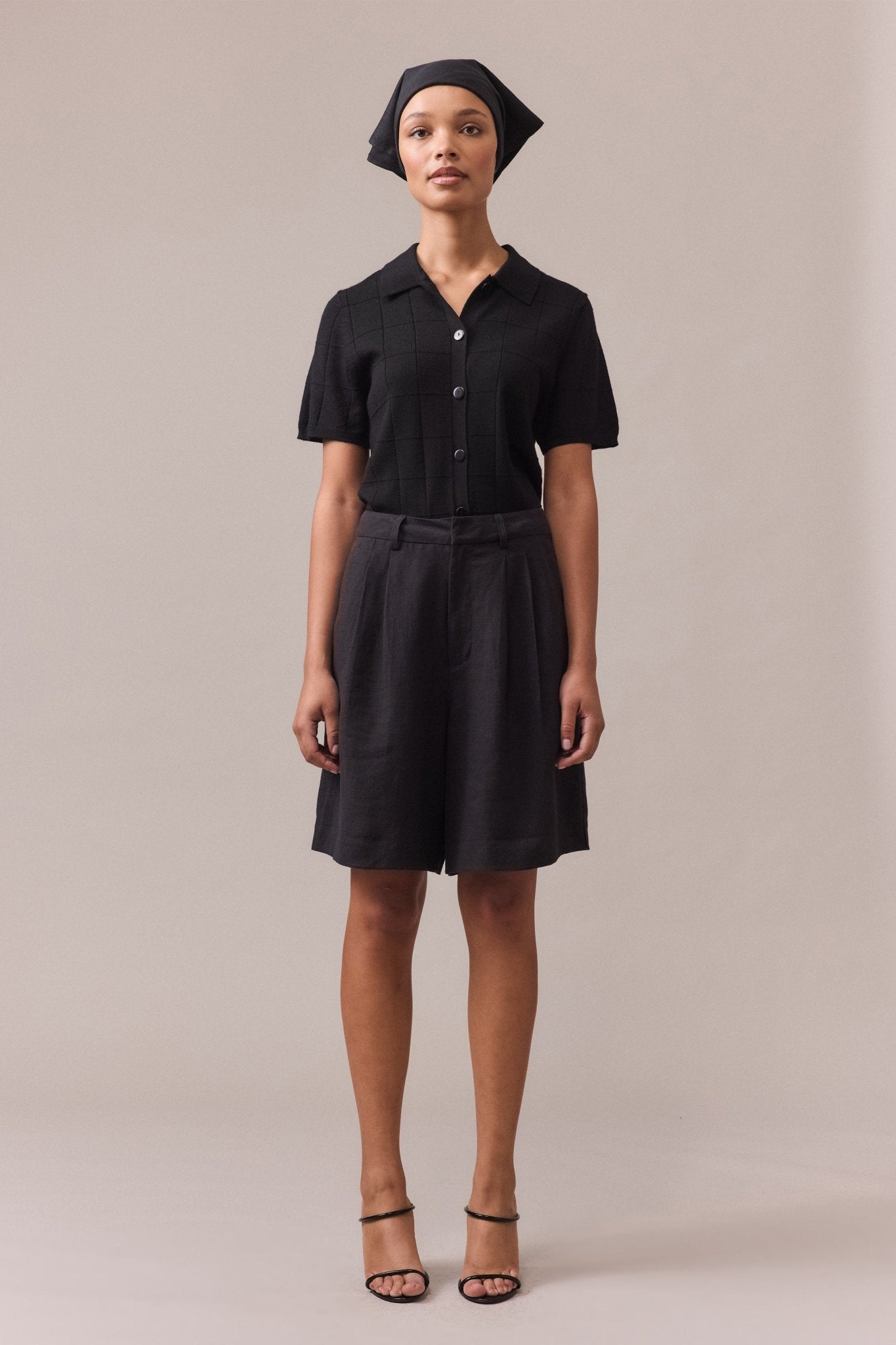 Woman wearing a black polo knit in 100% Merino Wool, standing with hands at her sides. The textured grid stitch, ribbed collar, and button-through placket are visible, highlighting the classic yet modern design.