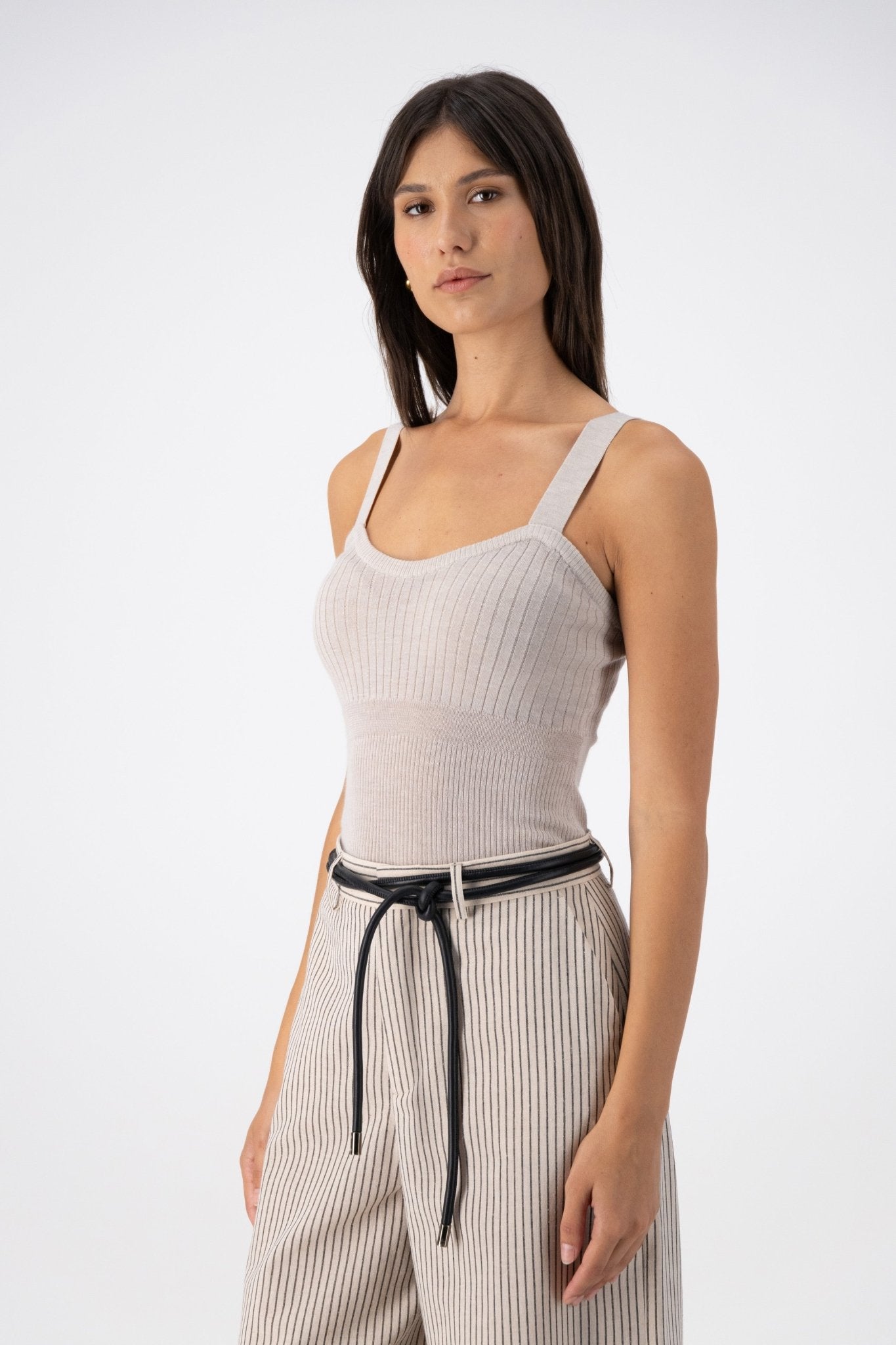 A woman is wearing a beige Low merino silk cashmere camisole paired with beige pants with black lines and leather belt wrapped around the waist.