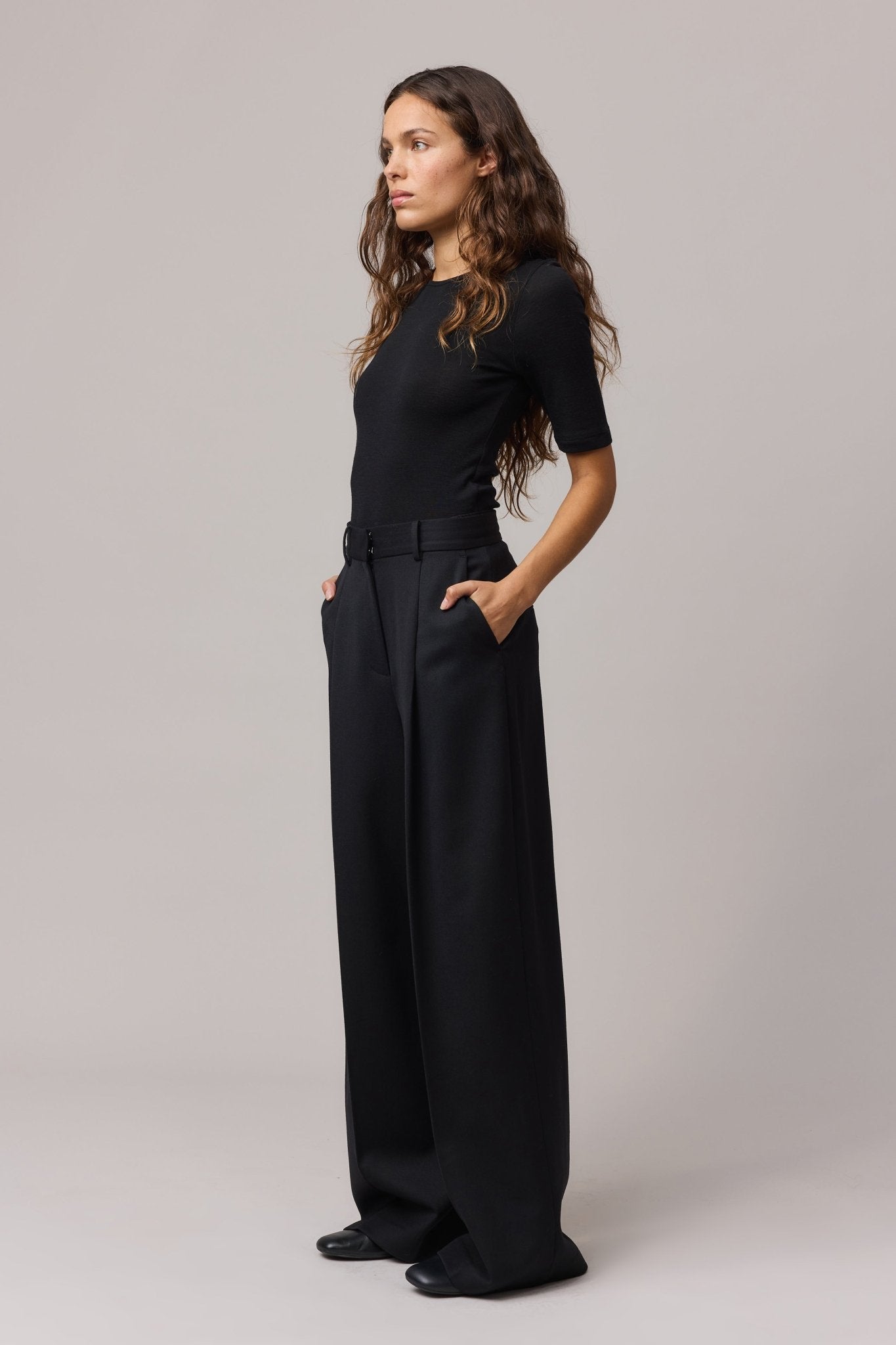 Side view of a woman wearing a black Merino Wool Crew Neck Tee with elbow sleeves and black linen pants with hands in the pockets.