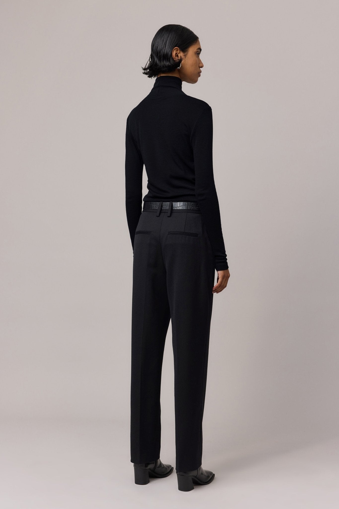 Back view of a woman wearing a black Merino Wool Slim Fit Skivvy with black pants.