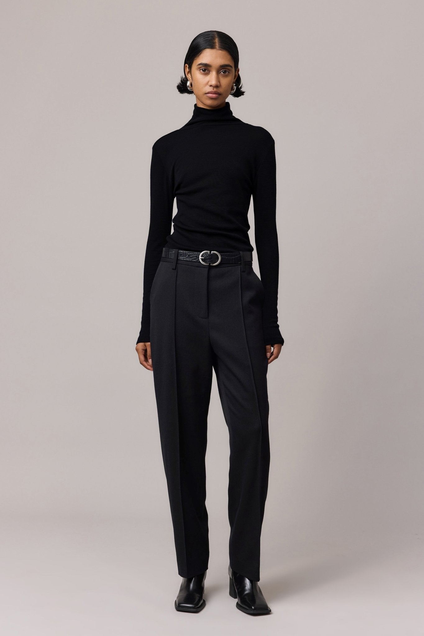 A woman styled in a black 100% Merino Wool Slim Fit Skivvy paired with sleek black pants. This cozy skivvy features a classic fitted neckline, offering both elegance and warmth.