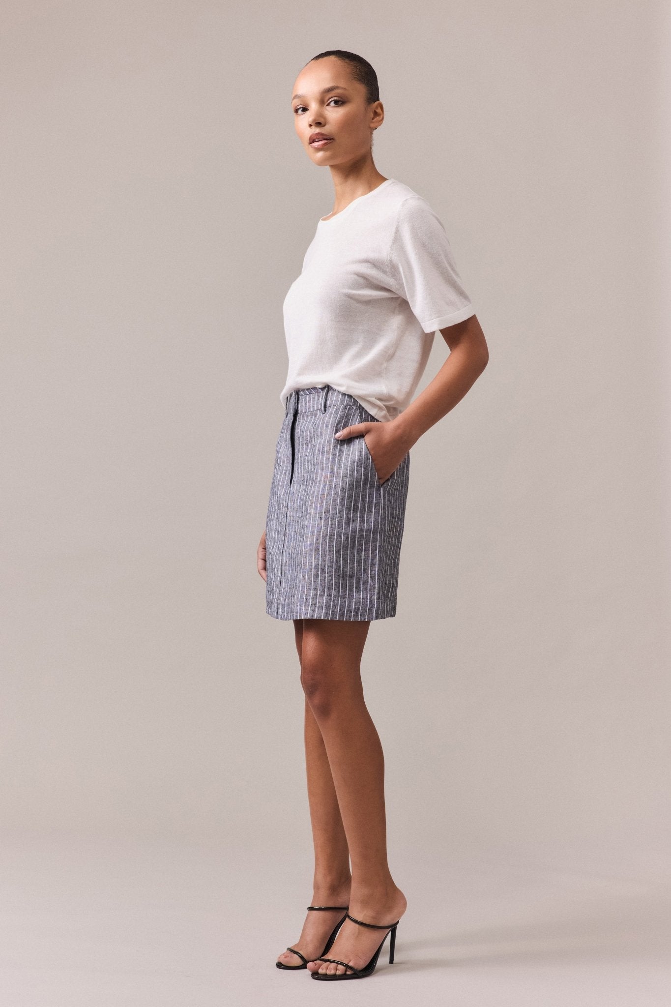 Woman wearing a white Nara Knit Tee with short sleeves, tucked into a grey mini skirt with white lines. She stands with one hand in her pocket, showcasing the relaxed fit of the tee.