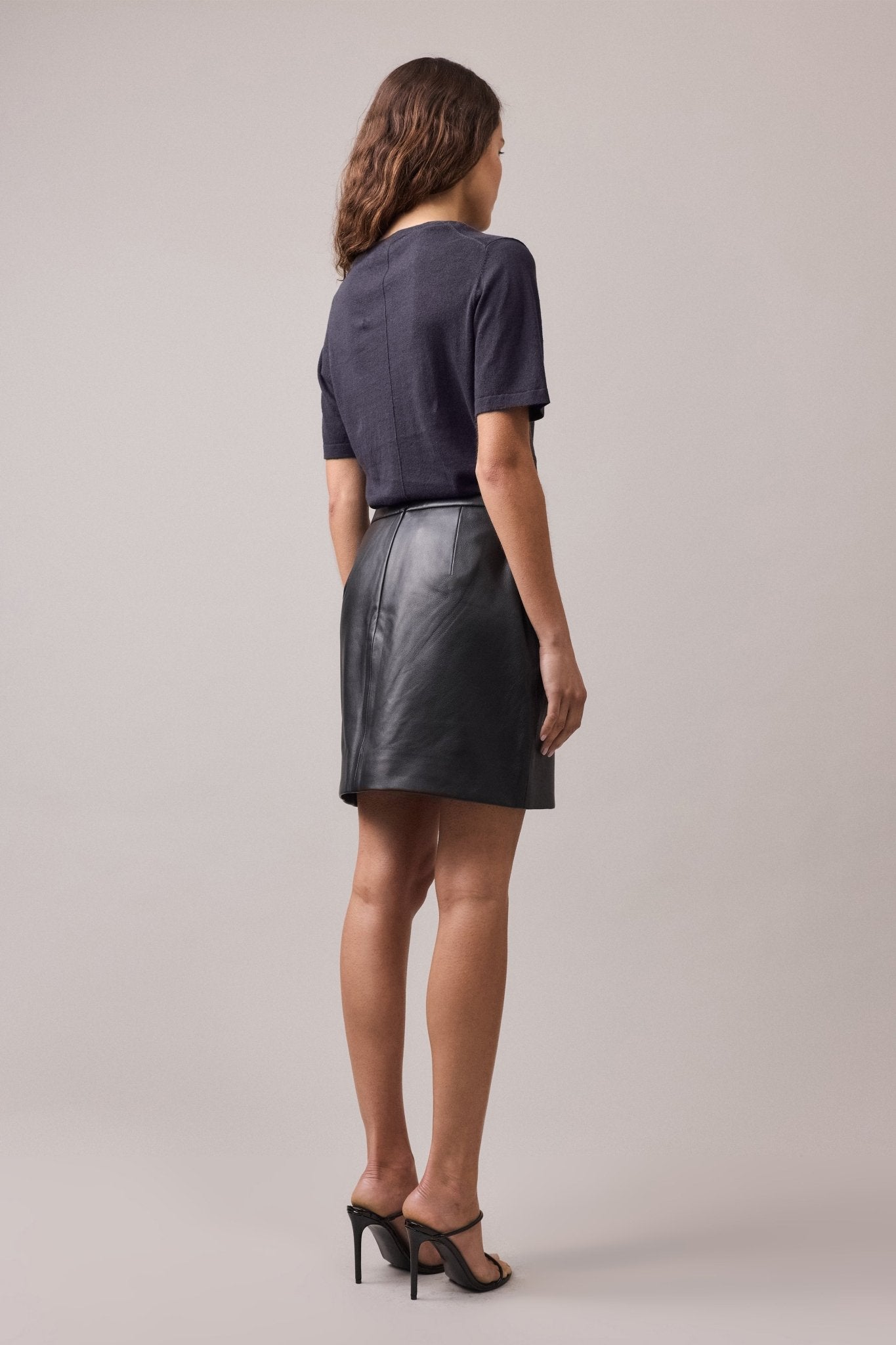 View of the woman from behind, wearing the navy Nara Knit Tee tucked into a mini leather skirt.