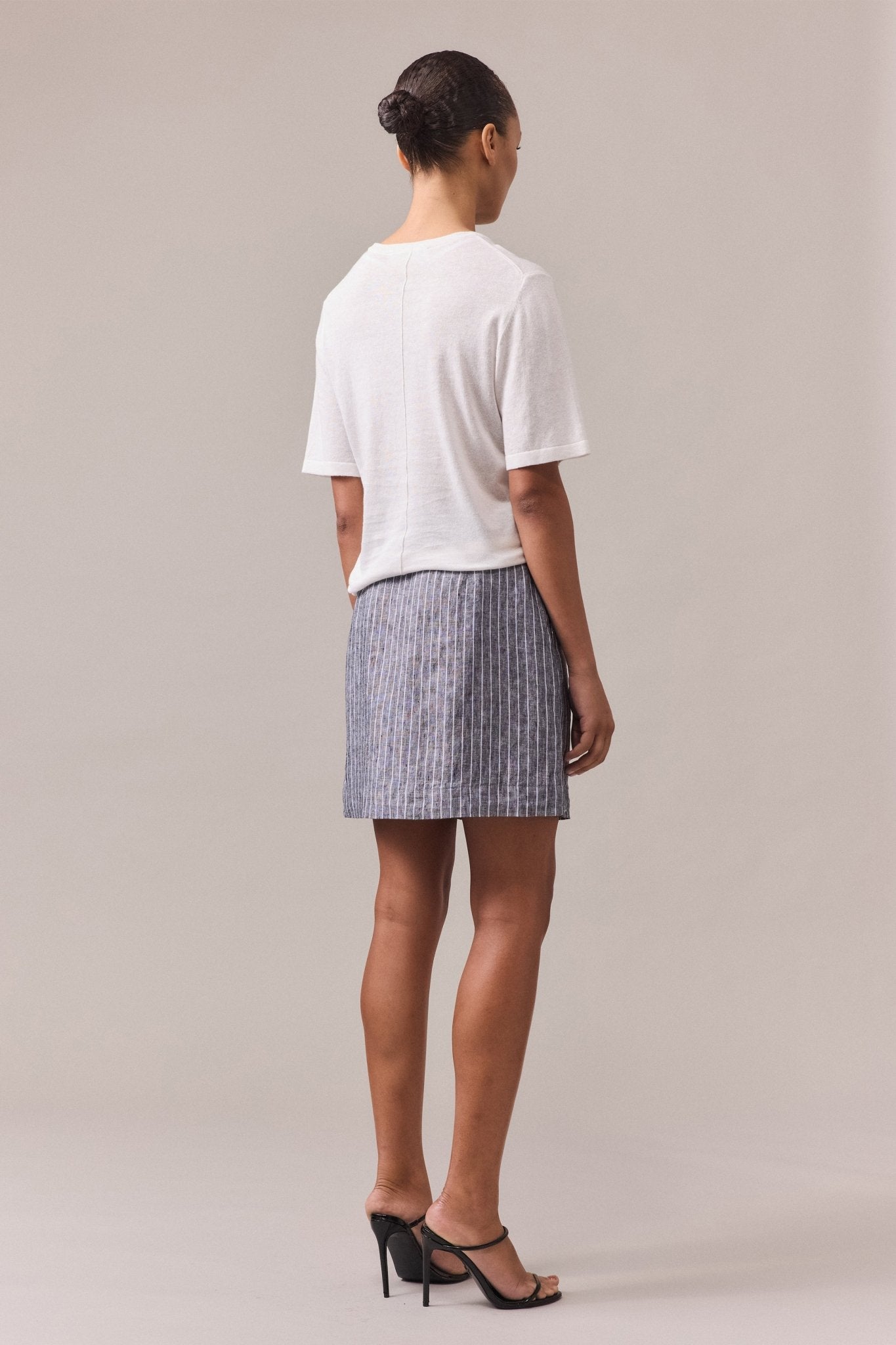 View of woman from behind, wearing the white Nara Knit Tee with short sleeves paired with a grey mini skirt with white lines.