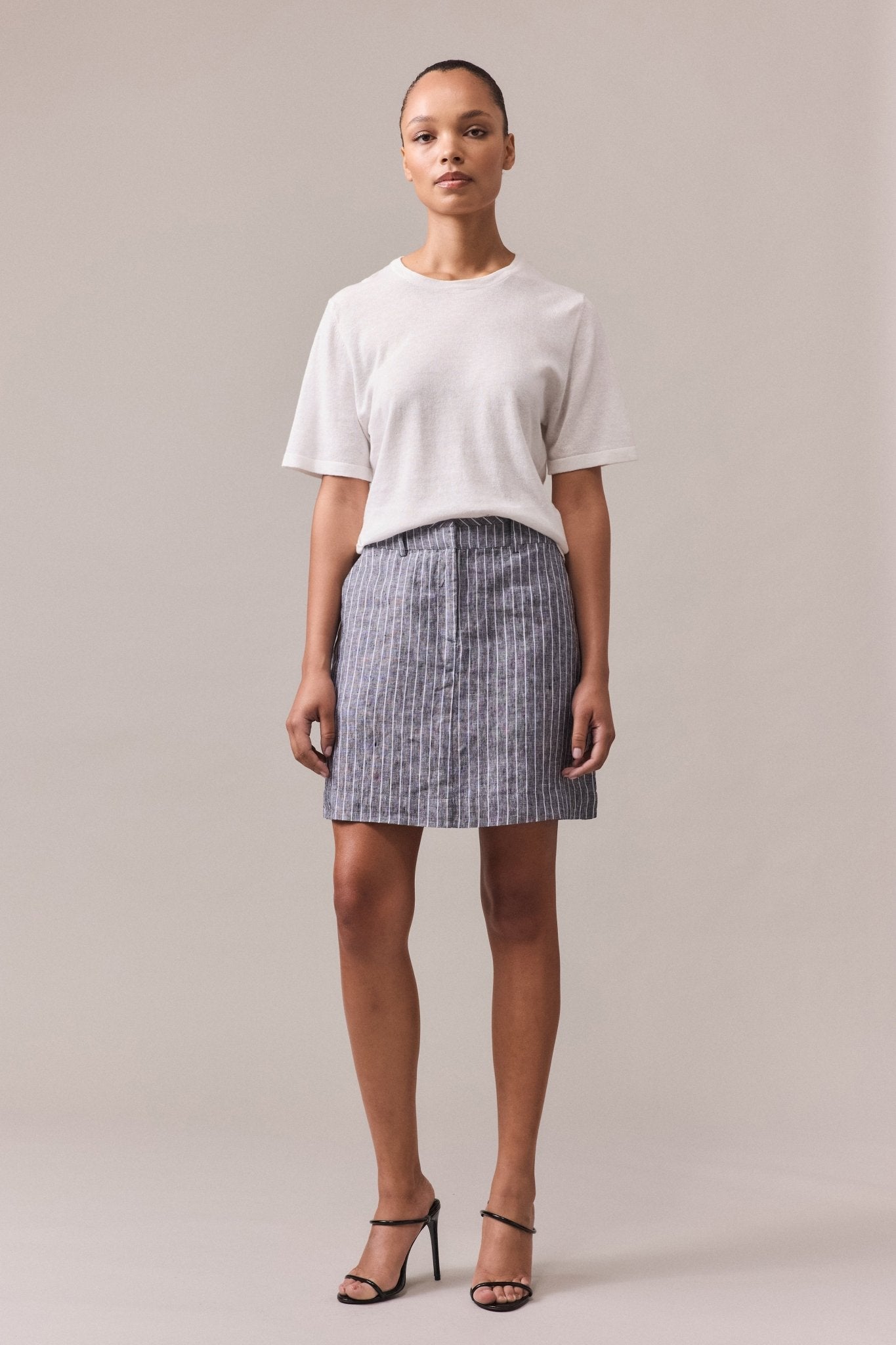 Woman wearing the Nara Knit Tee, a soft hemp and cotton blend, paired with a grey mini skirt with white lines. The tee features a relaxed fit and fine ribbed hem, collar, and cuffs, with linked knitwear seams.