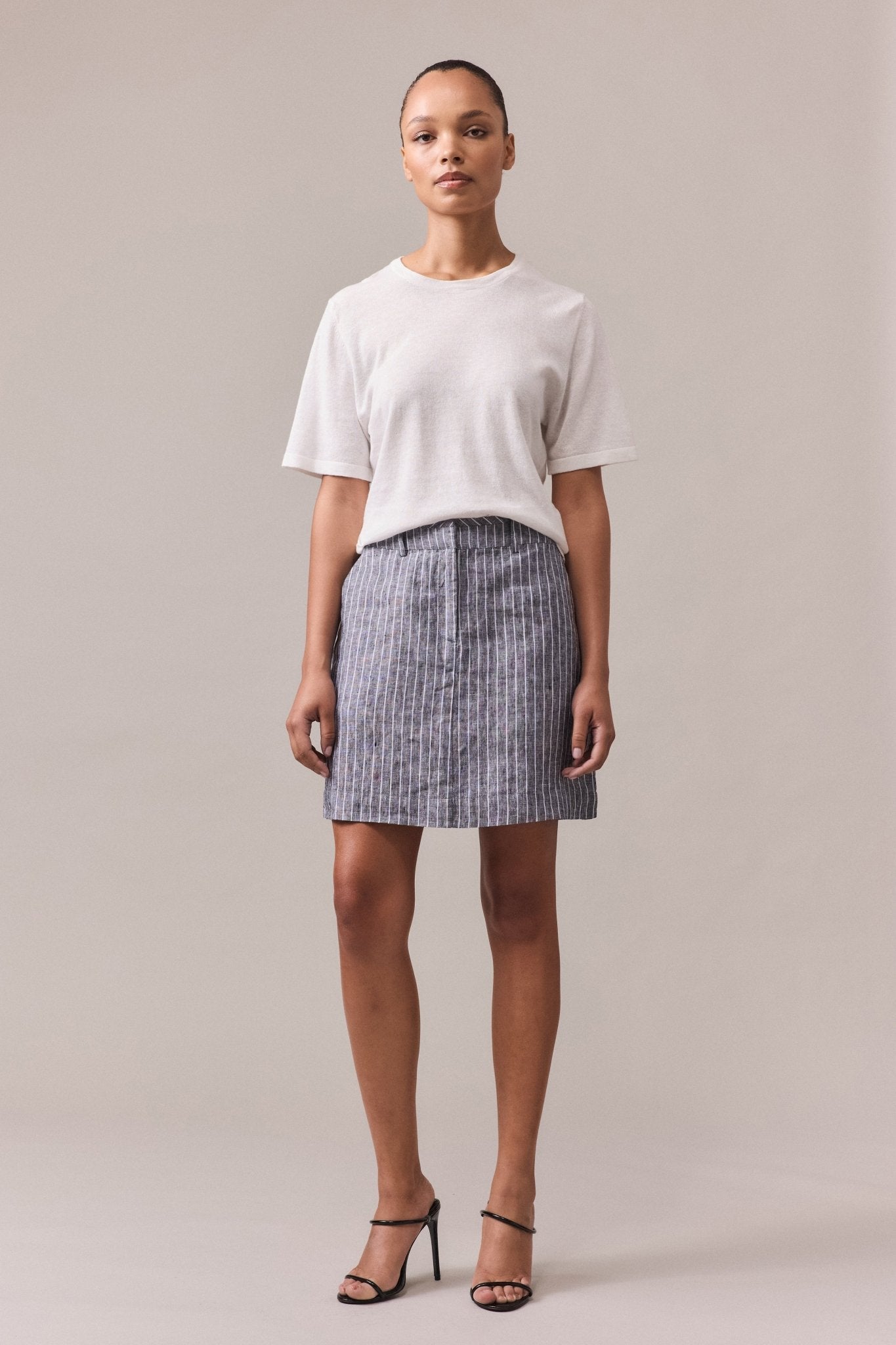 A woman wearing the Nara Knit Tee, a soft hemp and cotton blend, paired with a grey mini skirt with white lines. The tee features a relaxed fit and fine ribbed hem, collar, and cuffs, with linked knitwear seams.