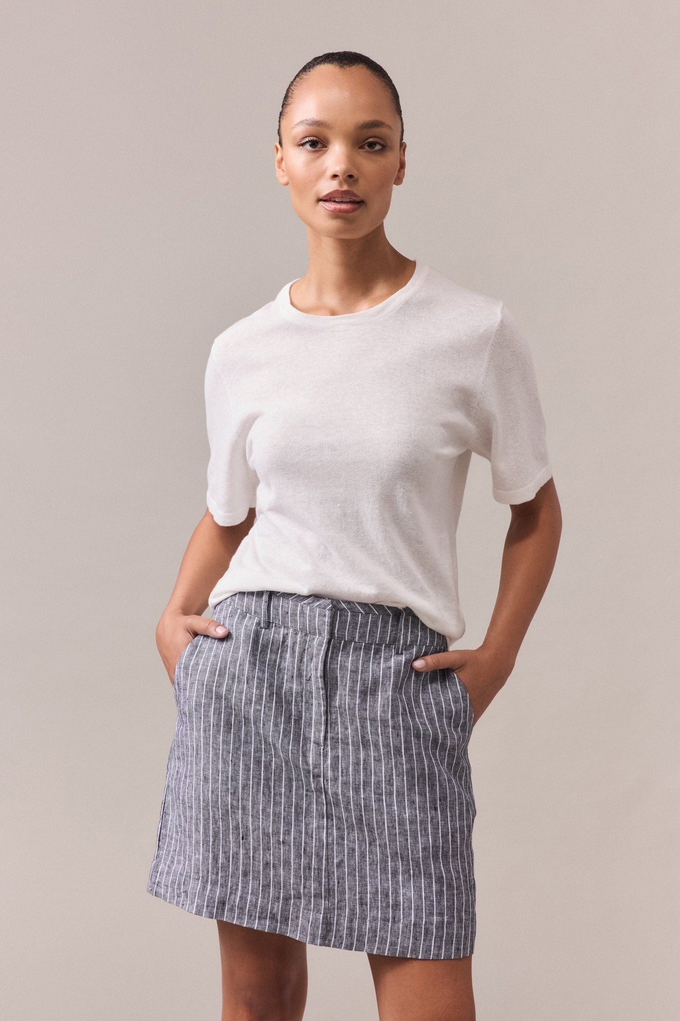 Woman wearing the Nara Knit Tee, a soft blend of hemp and cotton, paired with a grey mini skirt featuring white lines. She has her hands in the skirt pockets, showcasing the relaxed fit of the tee with fine ribbed hem, collar, and cuffs.